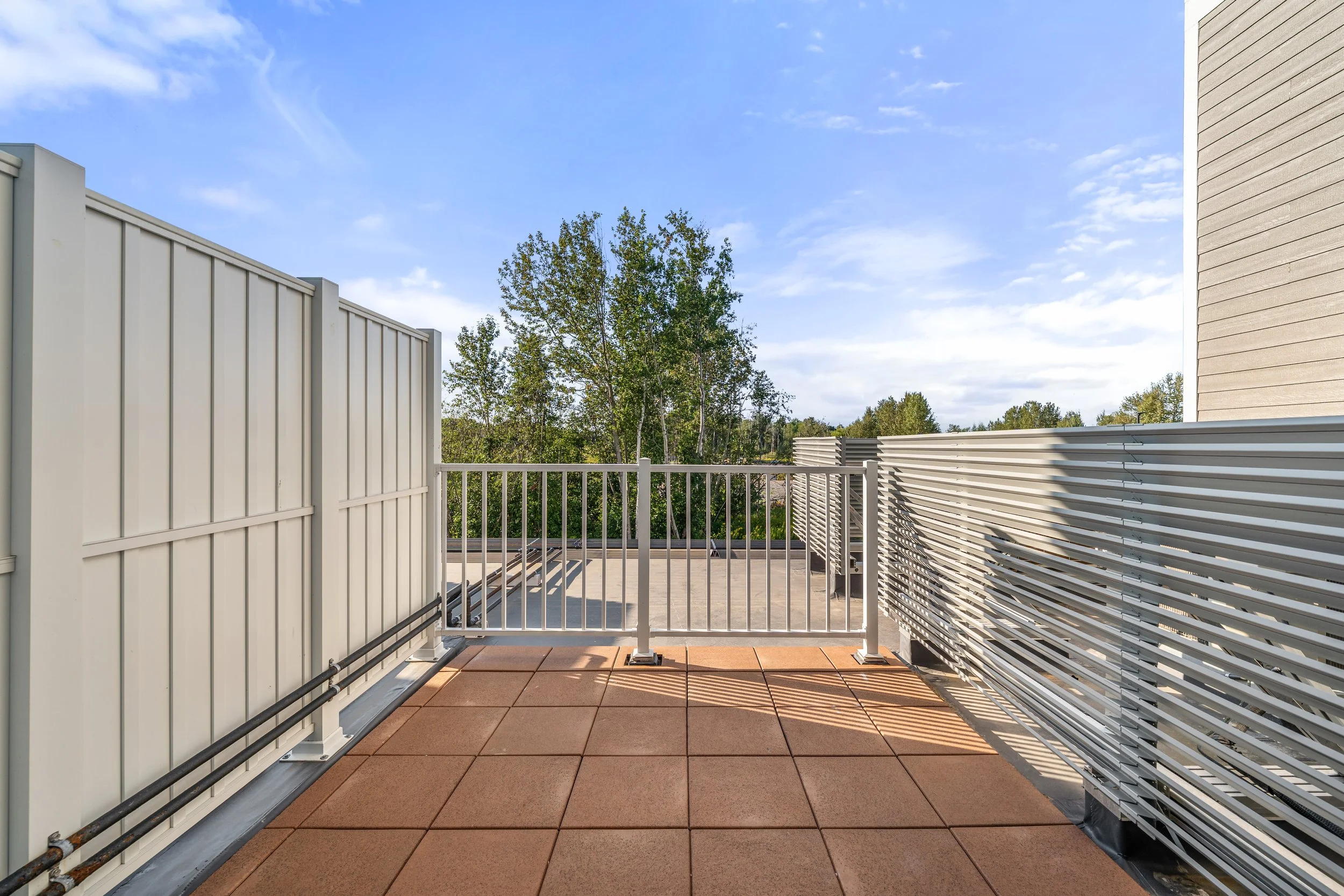 View from a balcony or rooftop with metal fences and railing, with a clear blue sky, some clouds, and trees in the background. The Residences at North Harbor on Lake Superior in Marquette Michigan.