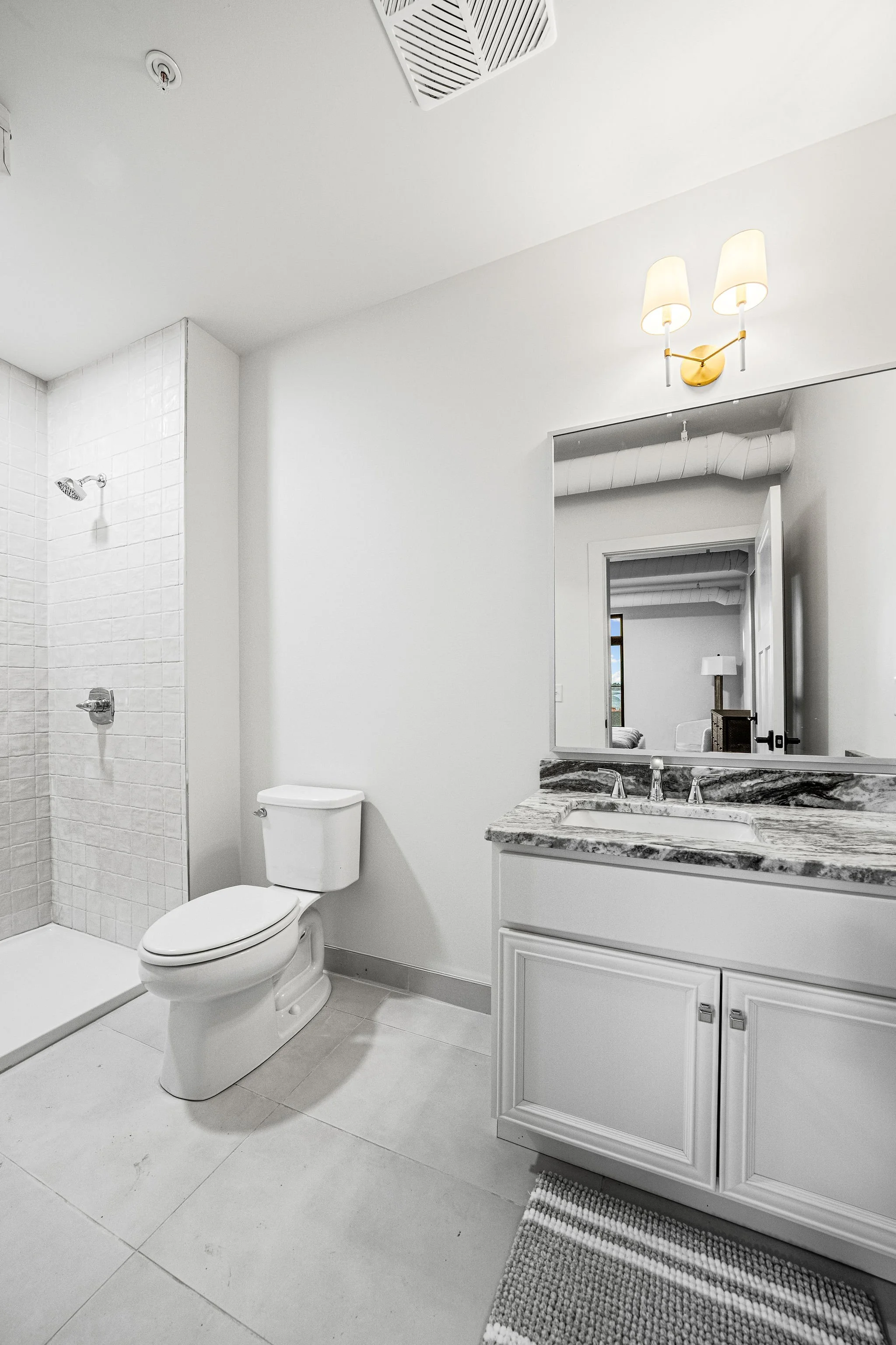 A modern bathroom with white walls, a shower area with tiled walls, a white toilet, a white vanity with a marble countertop and a built-in sink, a large mirror, a wall-mounted light fixture with two lamps, a small rug on the floor, and a reflection of a bedroom with a bed and a window in the mirror.