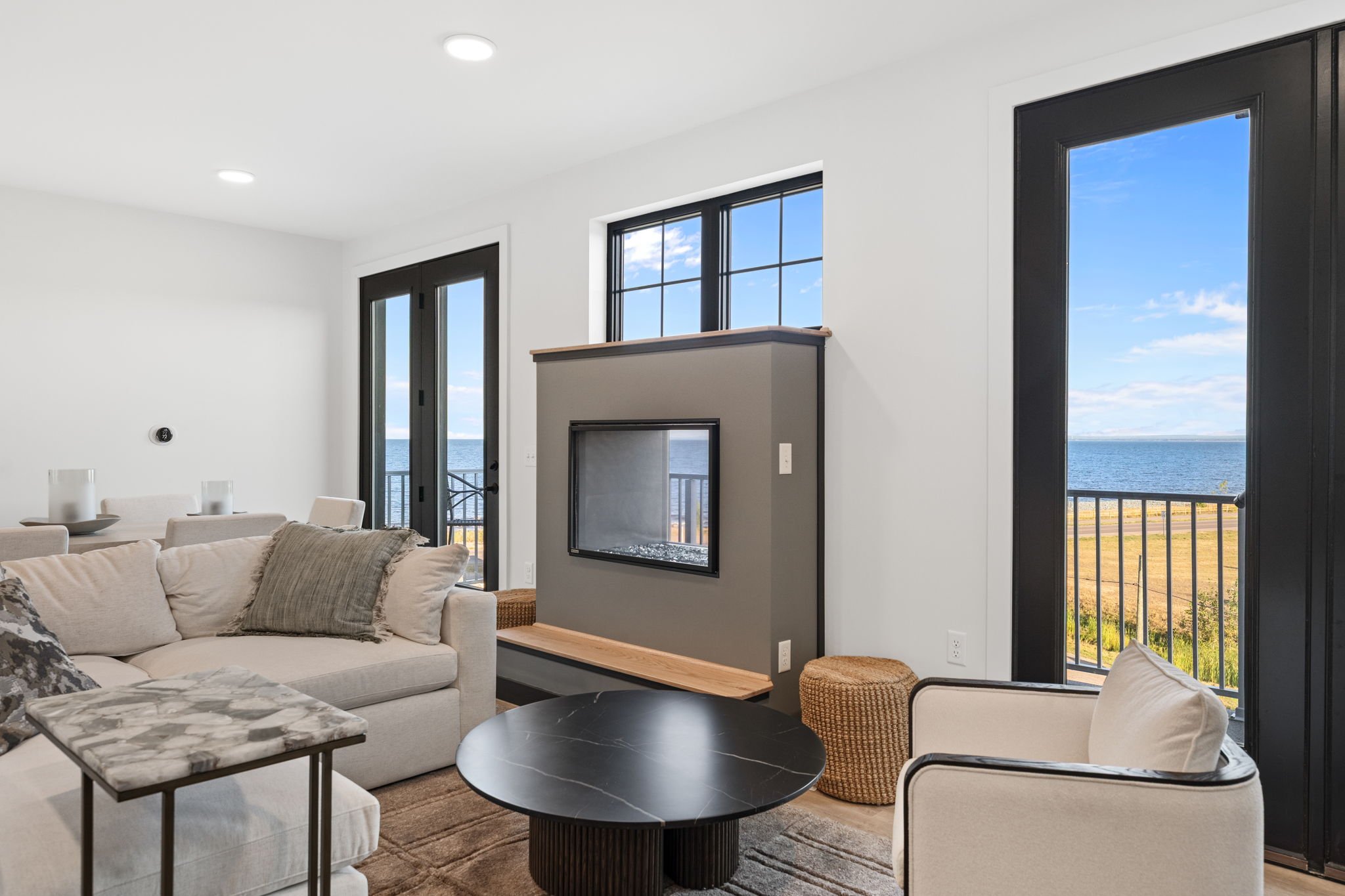Living room with a white sectional sofa, pillows, two black and white tables, a fireplace, and large windows with ocean views. The Residences at North Harbor on Lake Superior in Marquette Michigan.