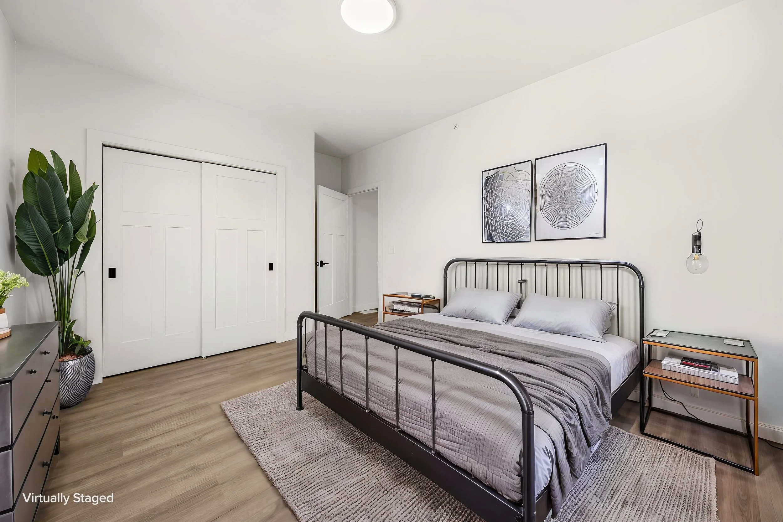 A bedroom with a black metal bed frame and gray bedding, a nightstand with books, a large green plant in a decorative pot, artwork with geometric designs on the wall. The Residences at North Harbor on Lake Superior in Marquette Michigan.