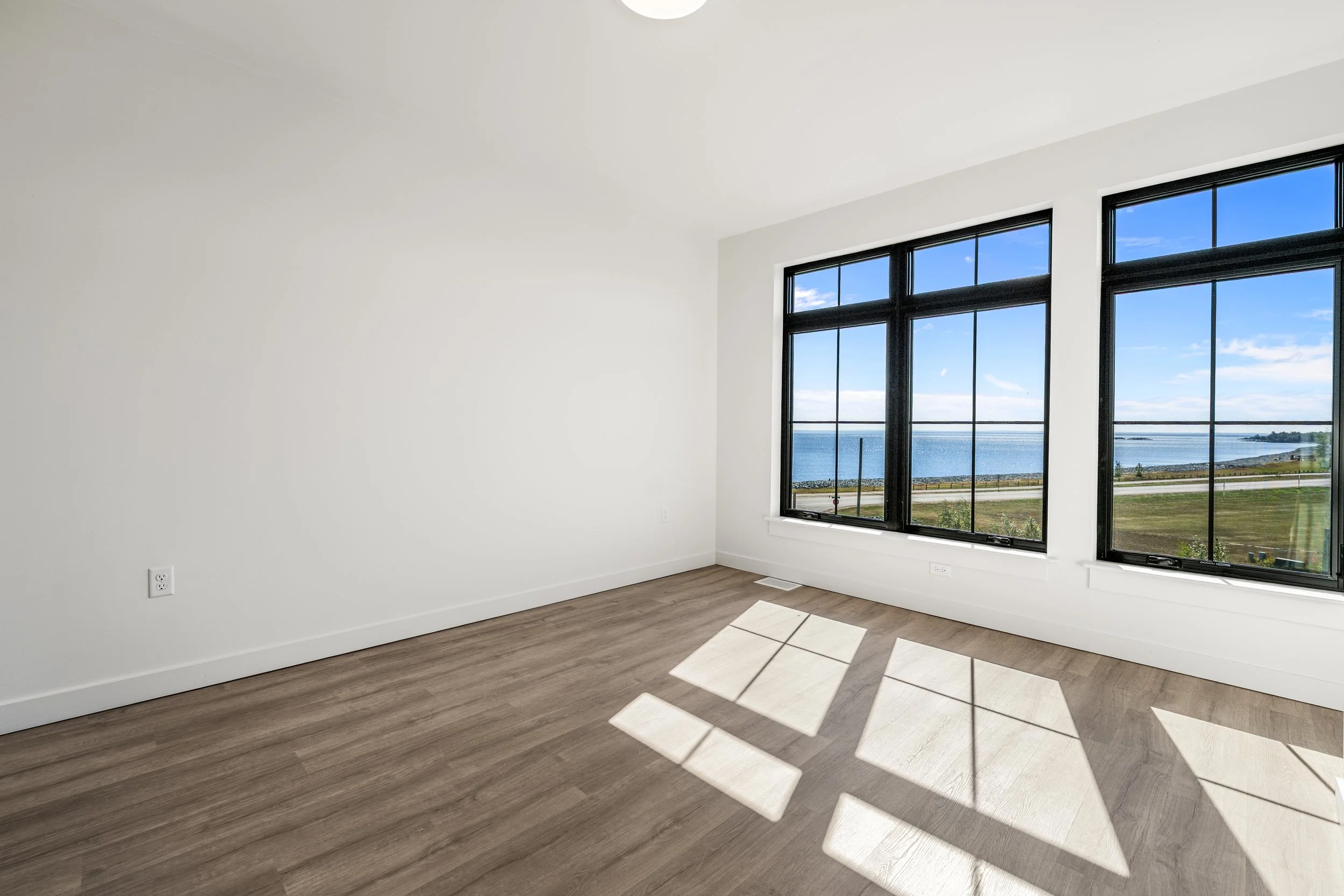 Empty room with large windows showing a view of water and sky, wood flooring, white walls, and sunlight casting shadows. The Residences at North Harbor on Lake Superior in Marquette Michigan.