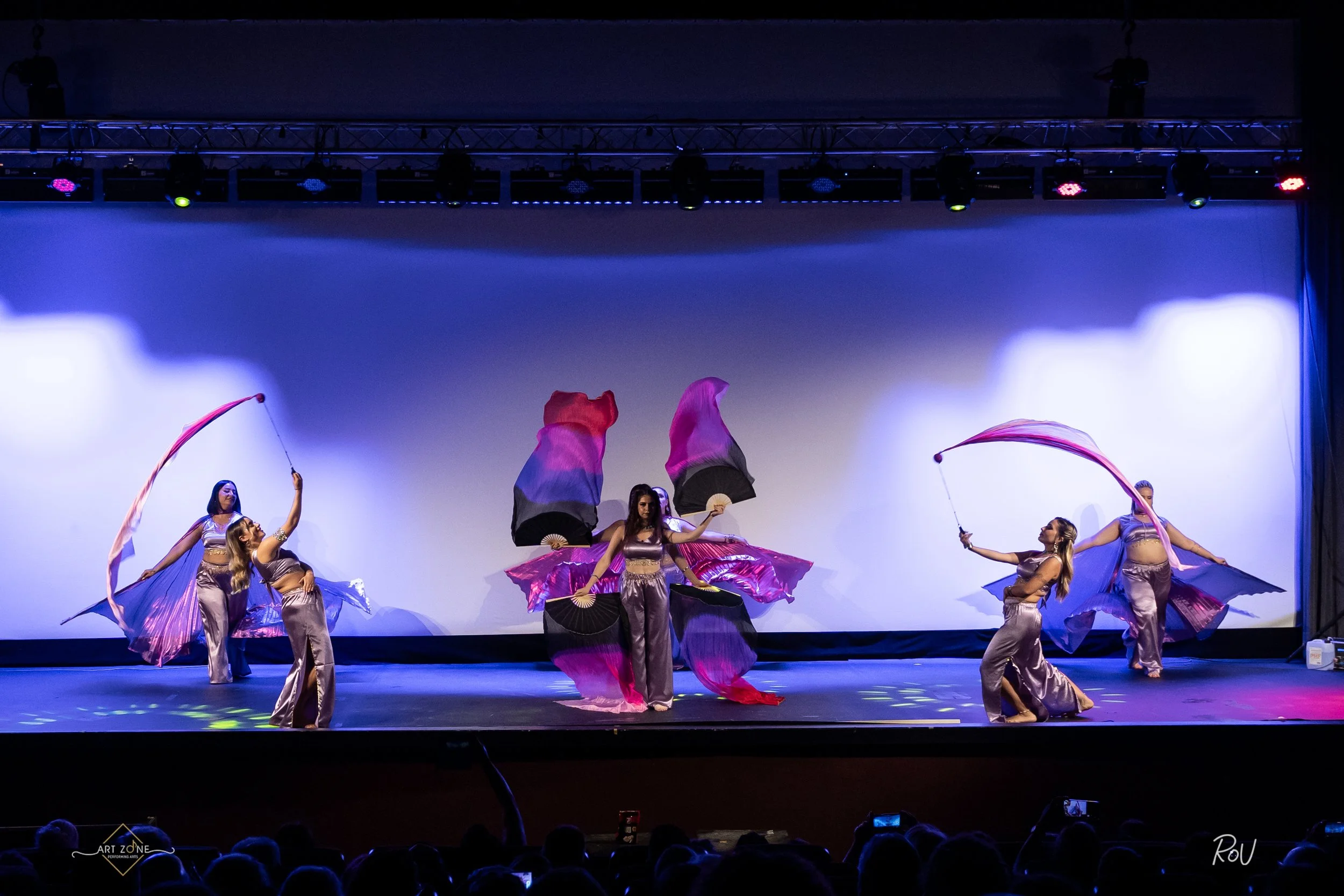 Group of five female dancers performing on stage, wearing black sparkly tops and blue sequined skirts, with blue background lighting.