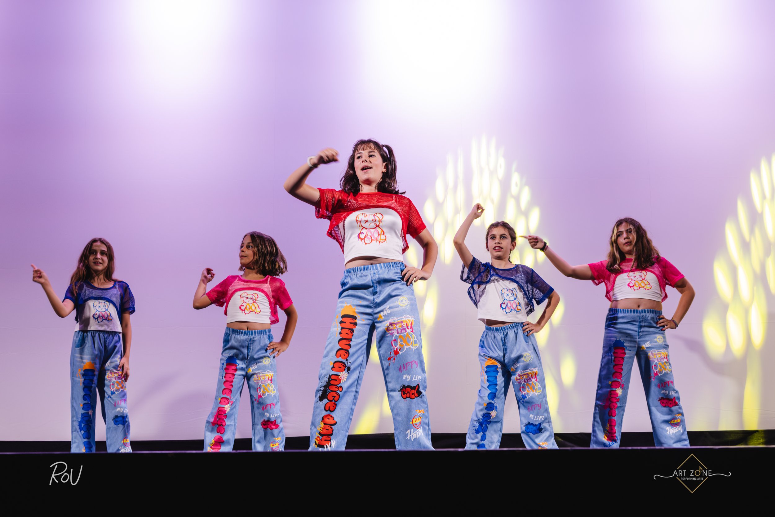 Four young women dance on stage, wearing casual clothing with bell-bottom jeans and crop tops, performing a synchronized dance routine.