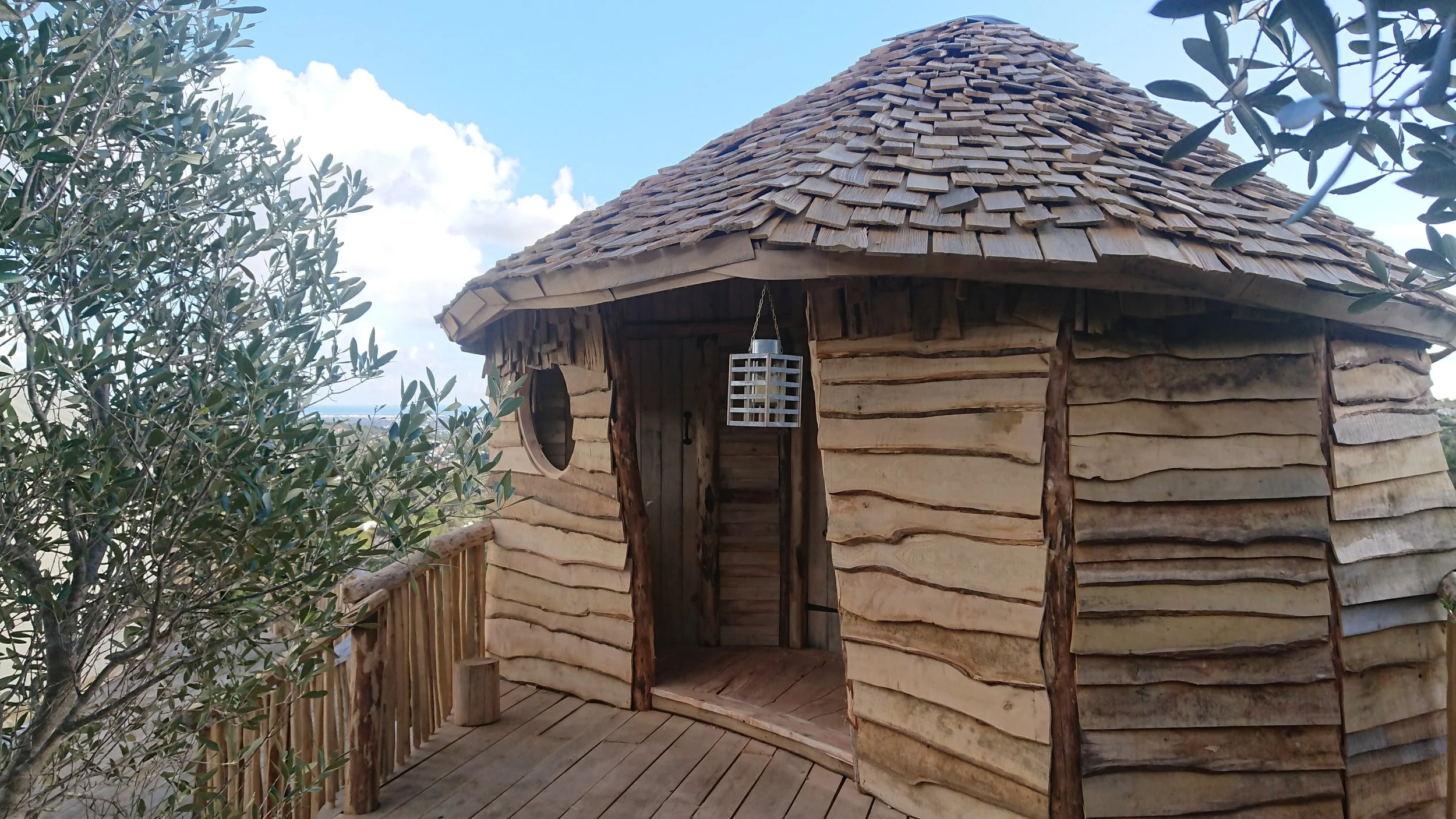 A small, round wooden treehouse with a shingled roof, a circular window, and a wooden door, surrounded by trees and open sky.