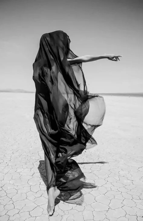 Person dressed in flowing black fabric standing on a cracked dry landscape, pointing into the distance.
