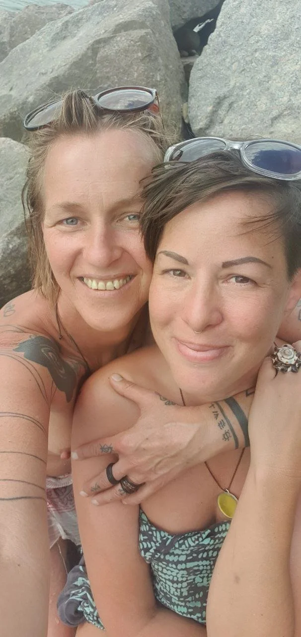 Two smiling women with short hair and tattoos taking a close-up selfie among rocks at a beach, wearing sunglasses on their heads.