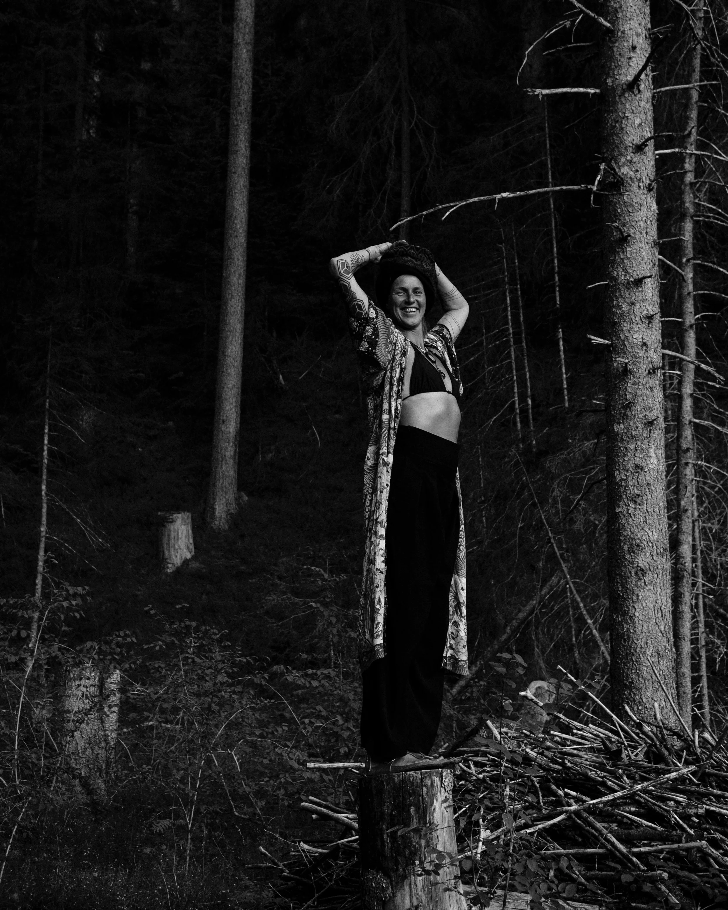 A woman standing on a tree stump in a forest, smiling and lifting her hands over her head.
