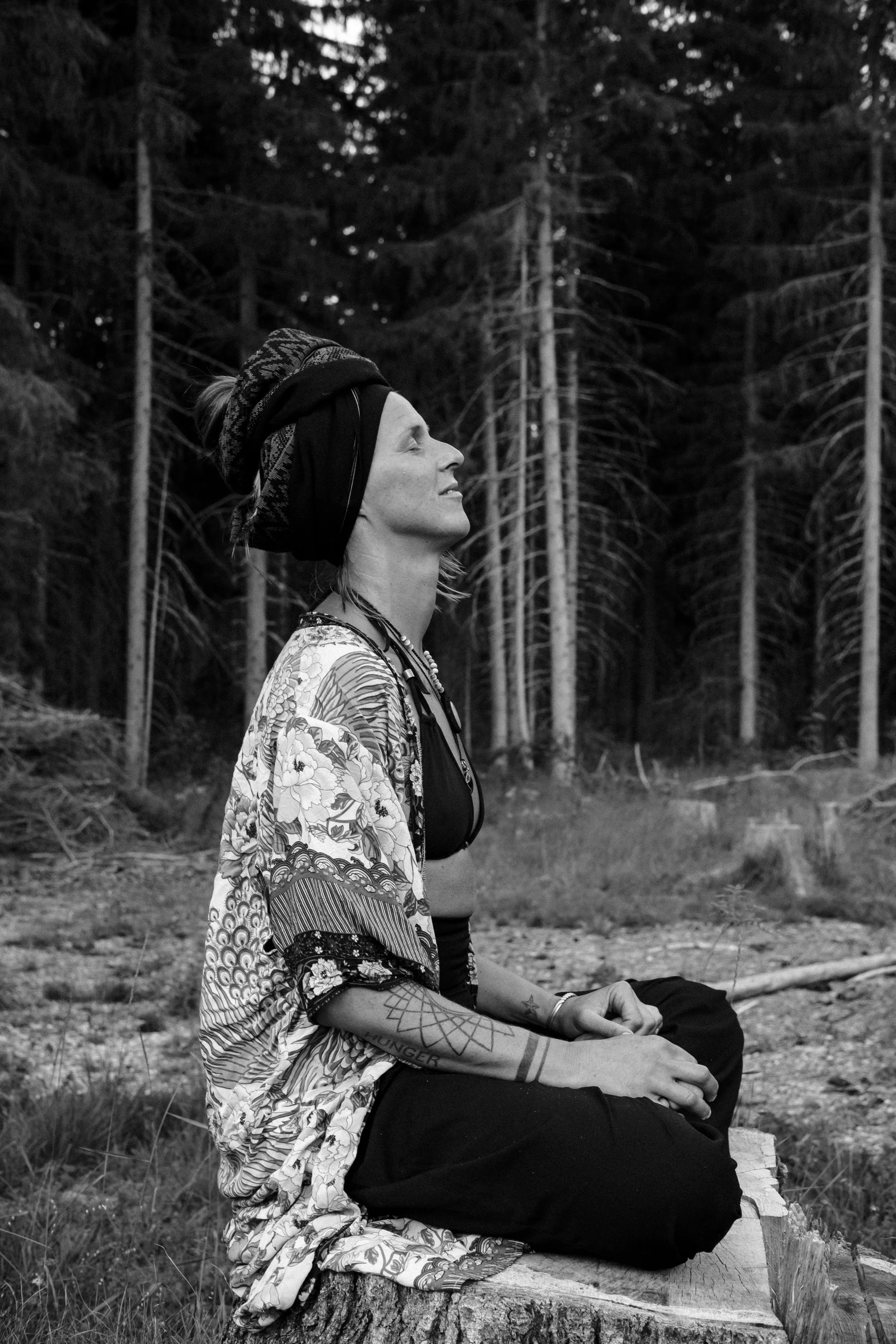 A woman sitting cross-legged on a log in a forest, wearing a patterned headscarf and loose clothing, practicing meditation with eyes closed.