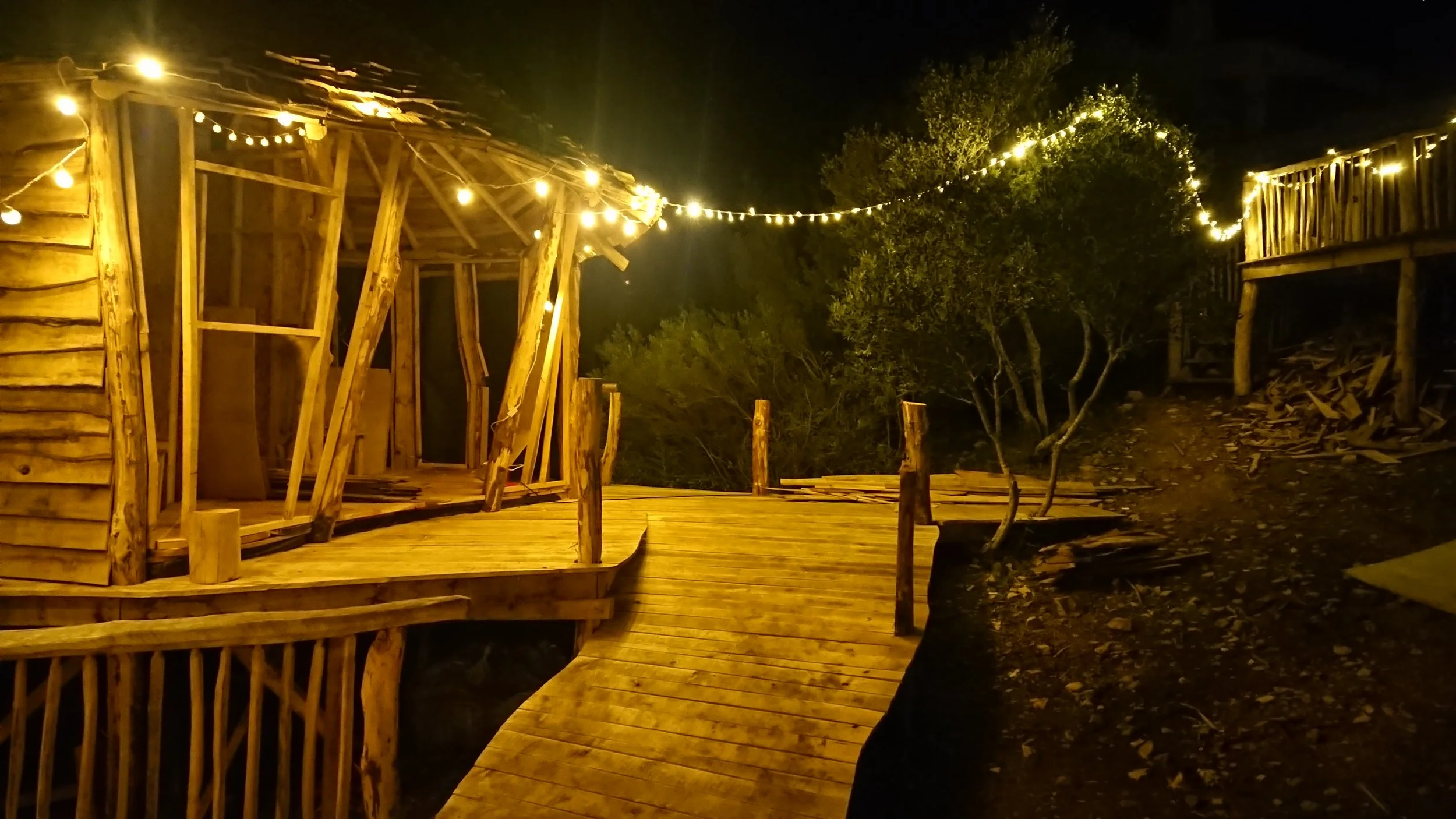 Wooden deck with string lights at night, under construction or renovation, with trees in the background.