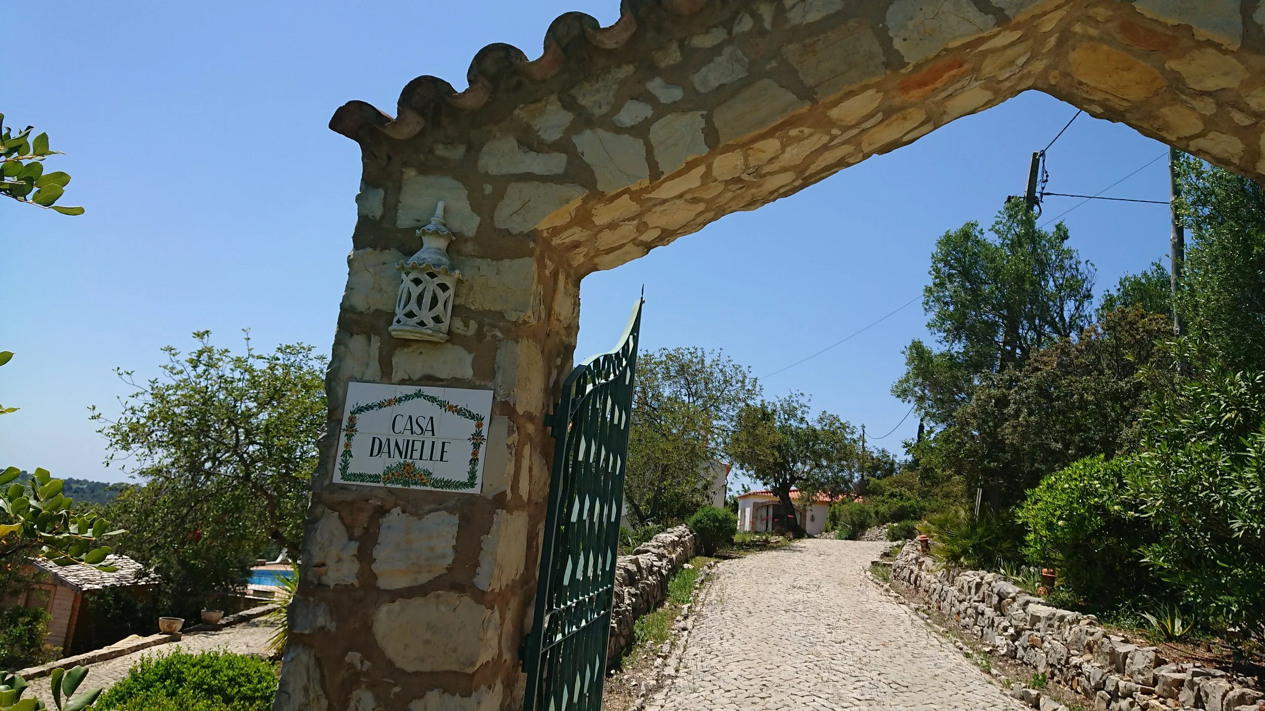 Stone archway with a sign that reads 'Casa Danielle' on a cobblestone path leading through a garden with trees and bushes, under a clear blue sky.