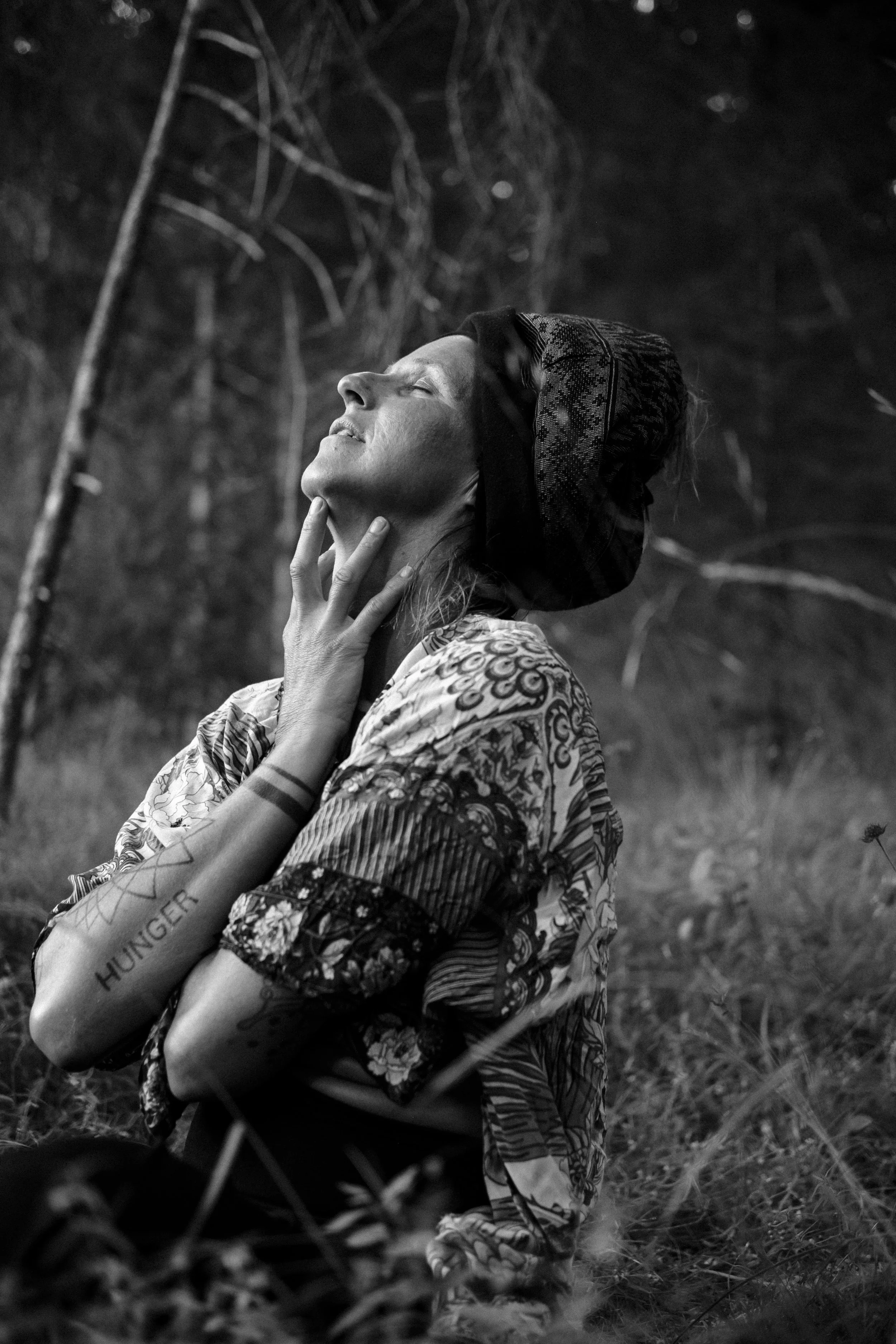 A woman wearing a patterned dress and a head covering kneels outdoors, eyes closed, with one hand touching her neck, seemingly in deep emotion or prayer. The setting appears natural, surrounded by tall grass and trees.