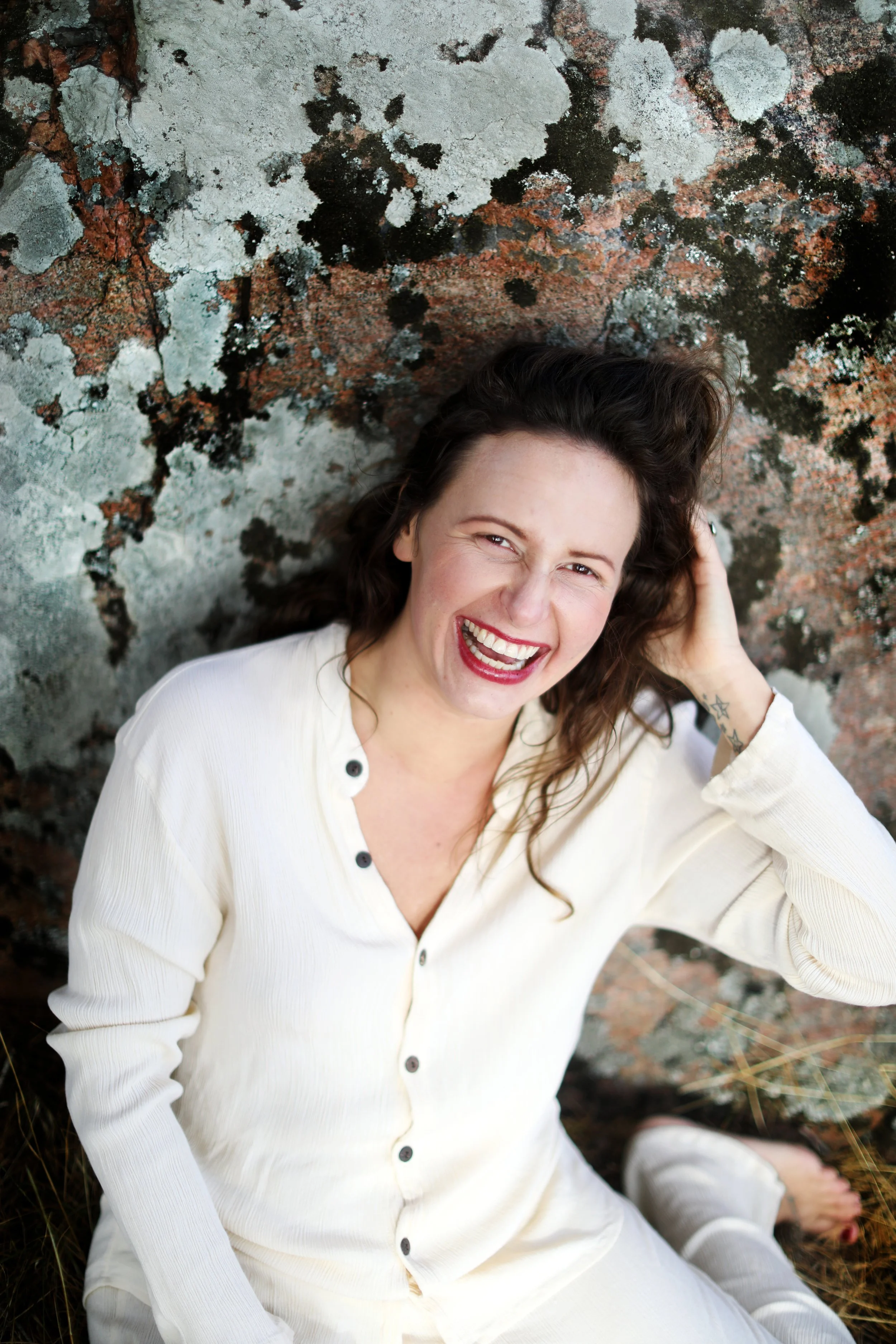 A woman with curly brown hair, wearing a cream-colored top, sitting in front of a textured, mossy rock wall, laughing with her head tilted back and hand in her hair.