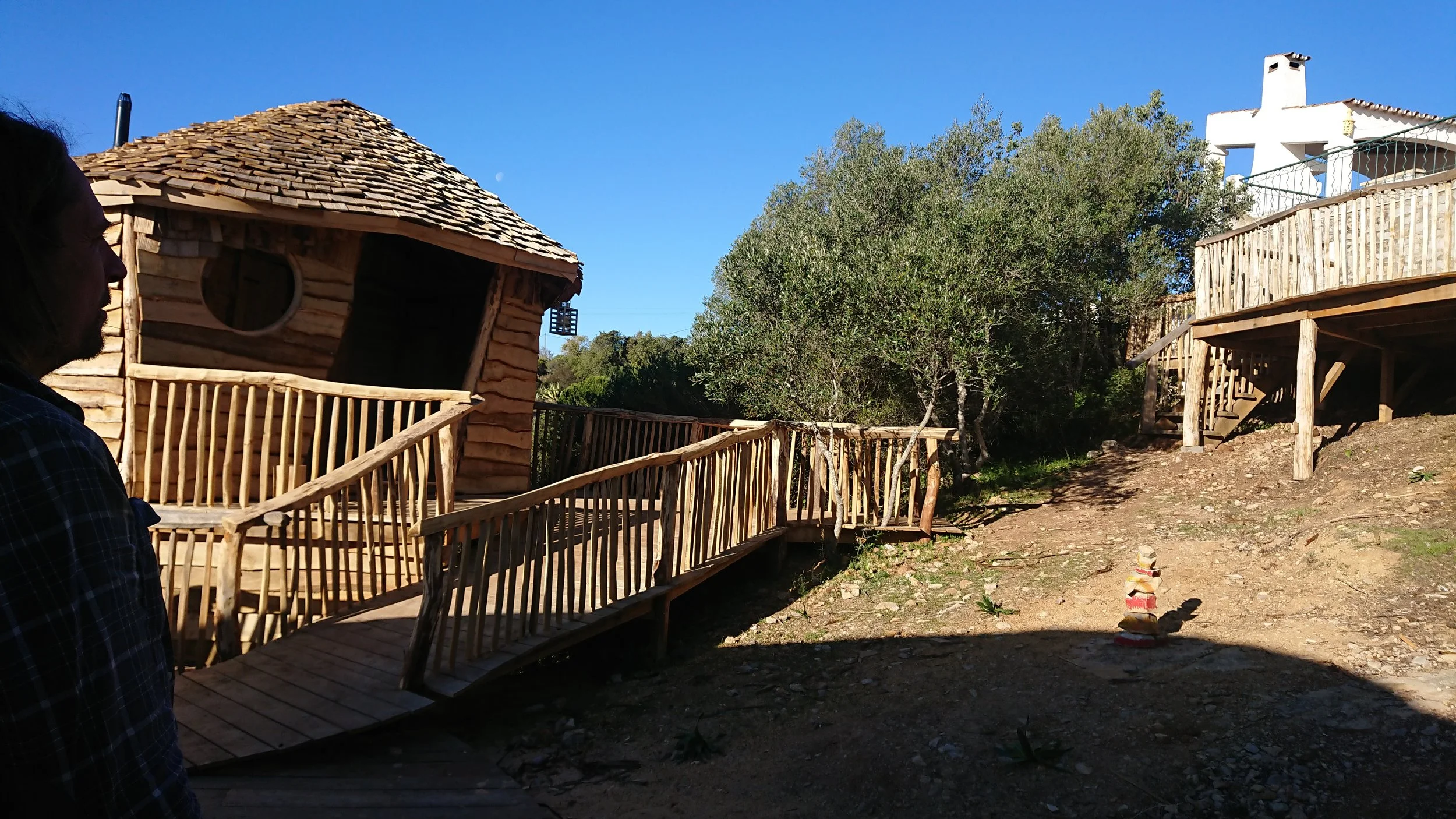 Person in plaid shirt standing near wooden treehouse on a hillside with trees and another house in the background under a clear blue sky.