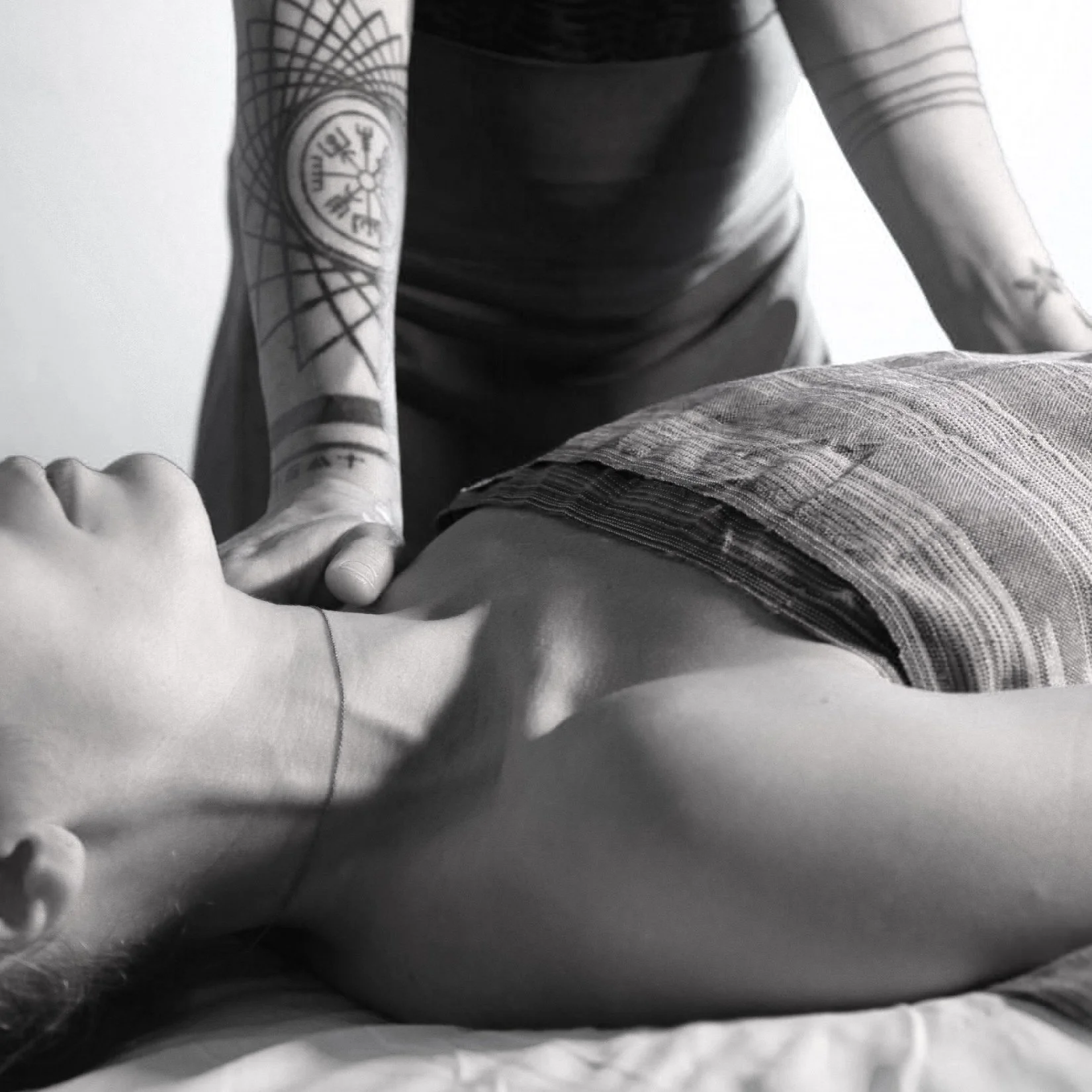 A person receiving a massage on their back, lying face up with a towel covering their lower body. The person giving the massage is leaning over and has tattoos on their arm.