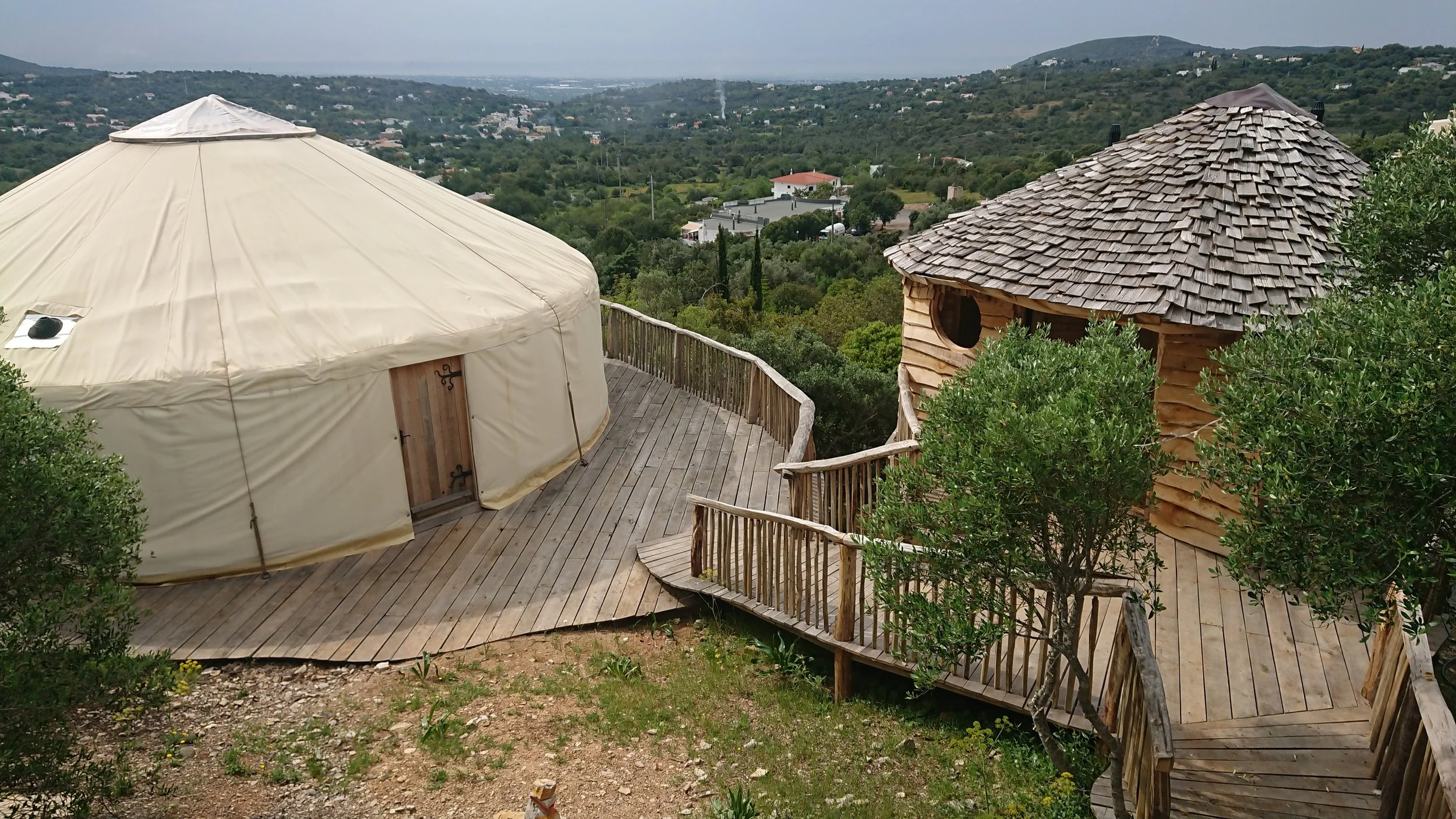 A wooden deck with a yurt on the left and a small wooden cabin with a shingled roof on the right, surrounded by green trees and overlooking a hilly landscape with scattered houses and distant mountains.