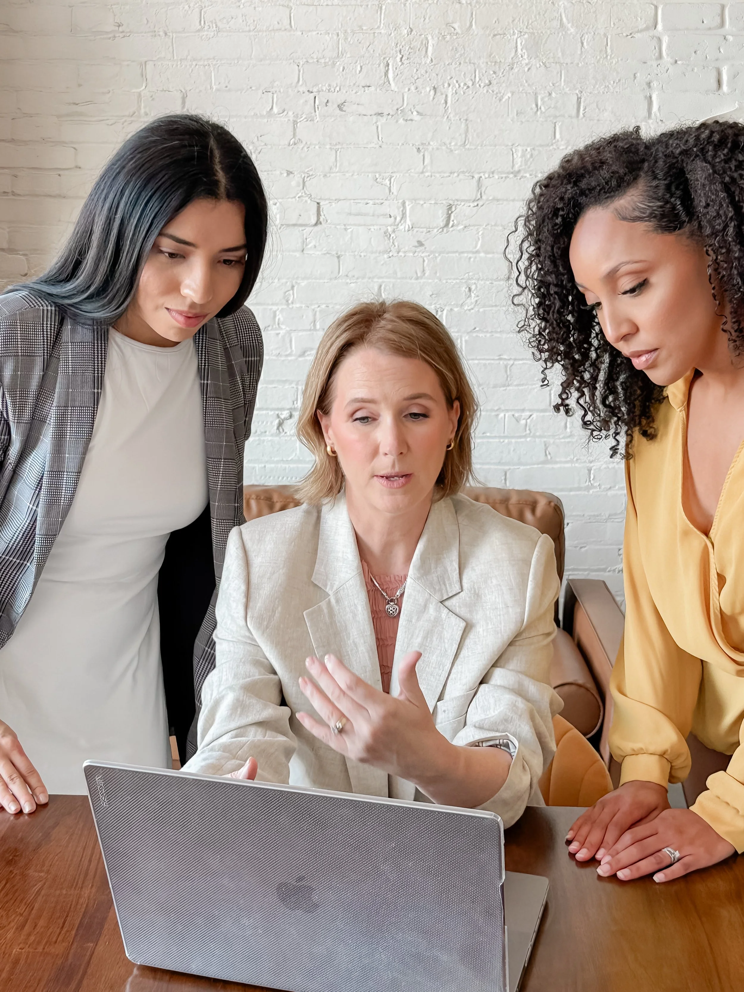 Three women gathered around a laptop, looking at the screen and discussing something, in an office with a white brick wall background.