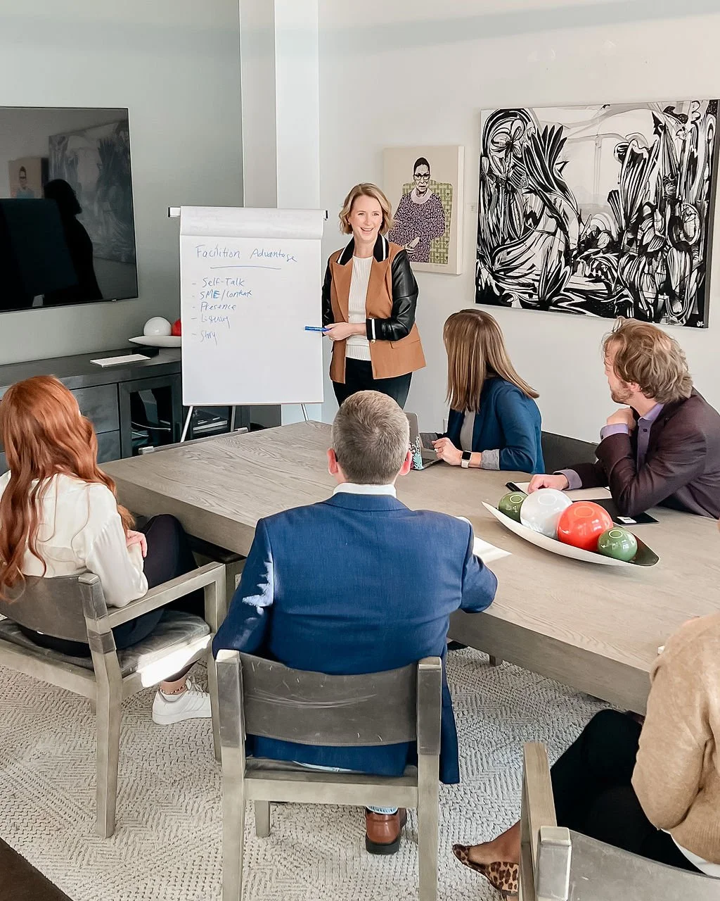 A woman presents in front of a whiteboard with notes, to a group of five seated people in a modern conference room. Artworks and a large TV are on the walls. The table has decorative balls.