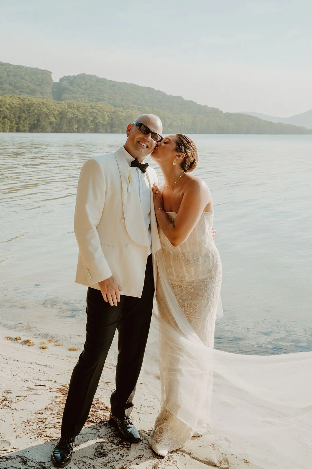 A woman in a white wedding dress and a man in a beige suit holding hands in a garden during sunset.