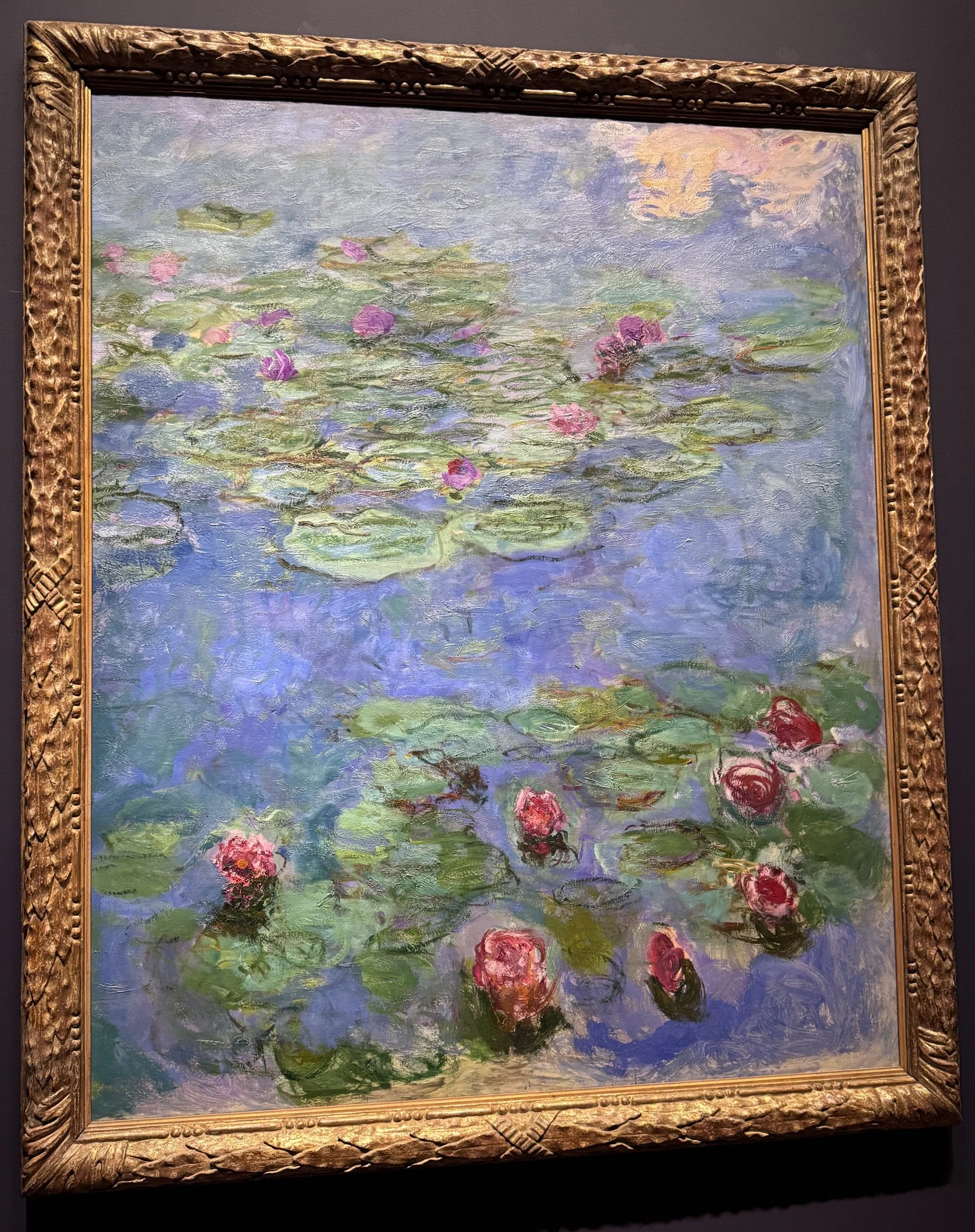Painted after Monet's sojourn to Venice, this water lily painting features loosely painted pink and red lilies, with a soft cloud reflecting on the water at the top of the painting.
