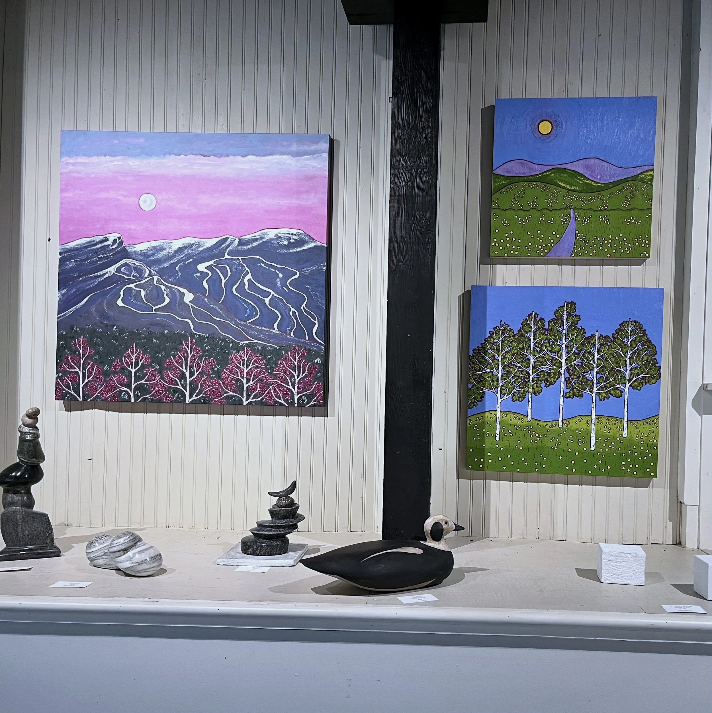 Several abstract paintings displayed on a wall and sculptures on a table in a gallery setting.