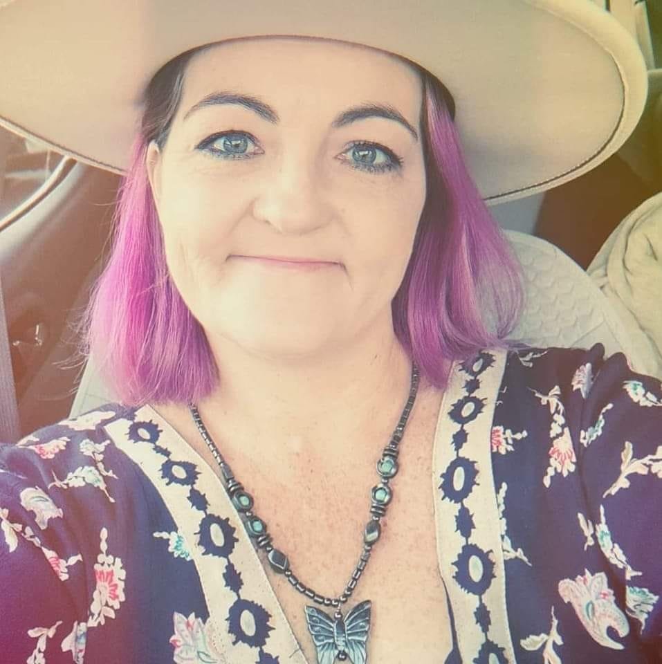 Woman with purple hair in a floral top and wide-brimmed hat, smiling in a car.