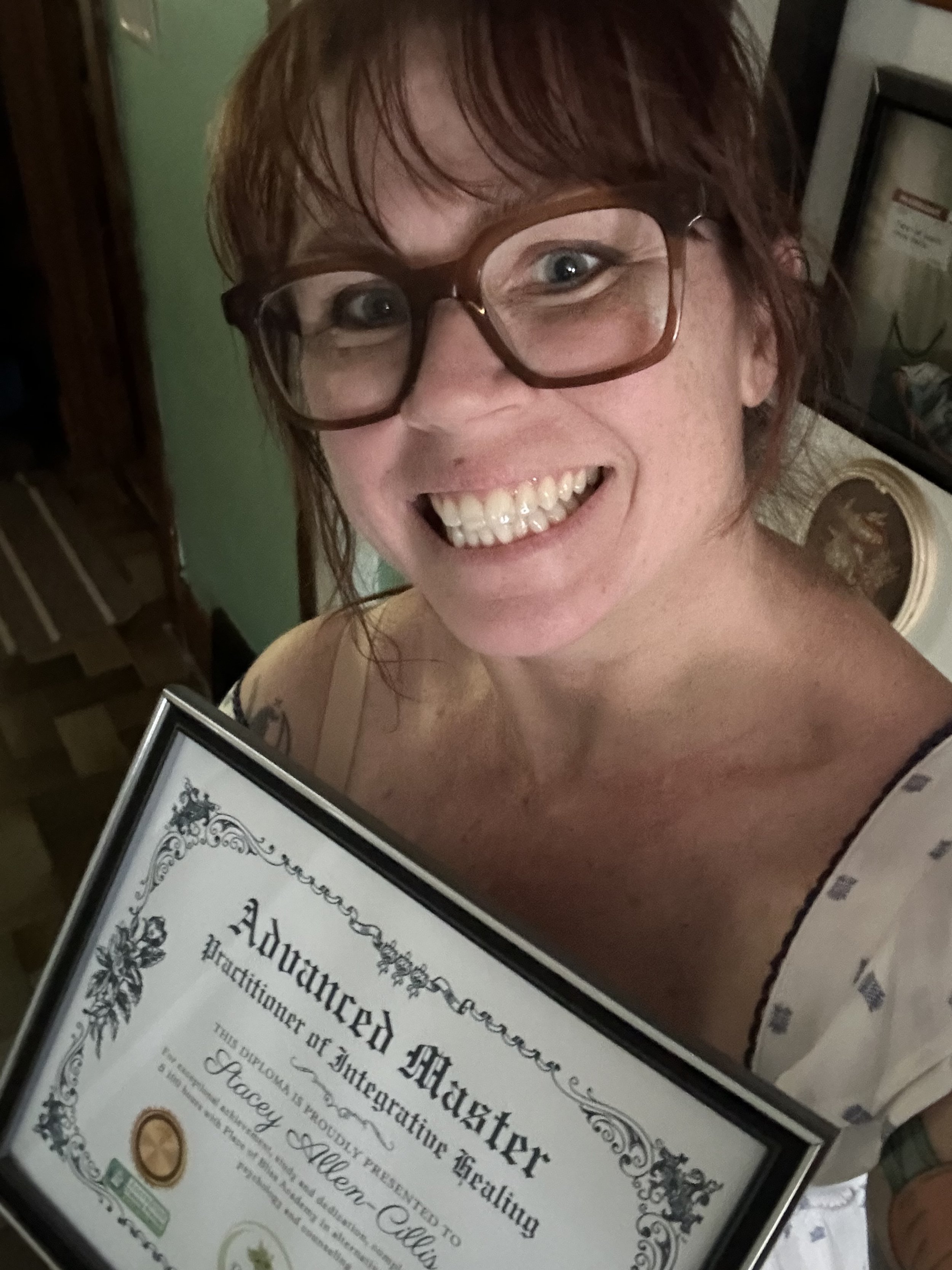 A smiling person wearing glasses holding a framed diploma titled 'Advanced Master Practitioner of Integrative Healing.' The diploma is partially visible in the image.