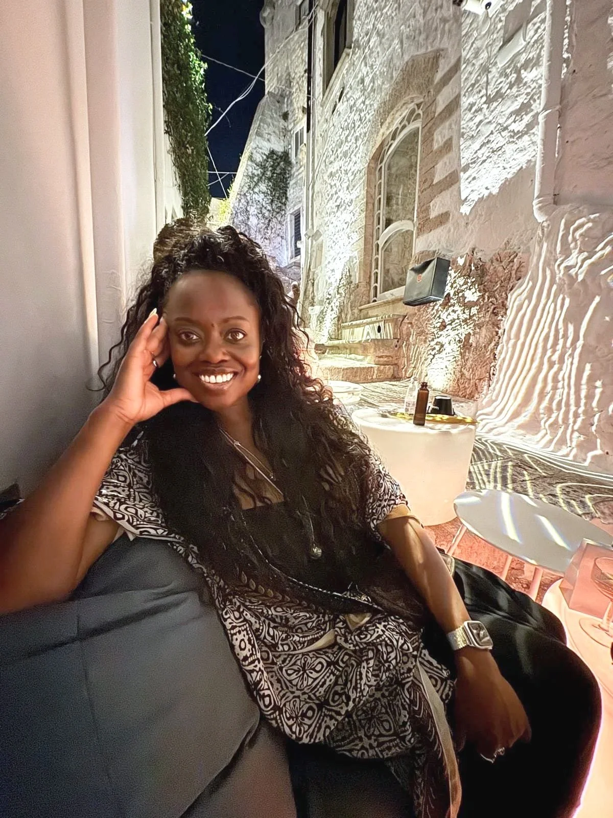 A woman with long curly hair smiling and sitting on a couch at night in an outdoor restaurant or café with stone walls and balconies in the background.