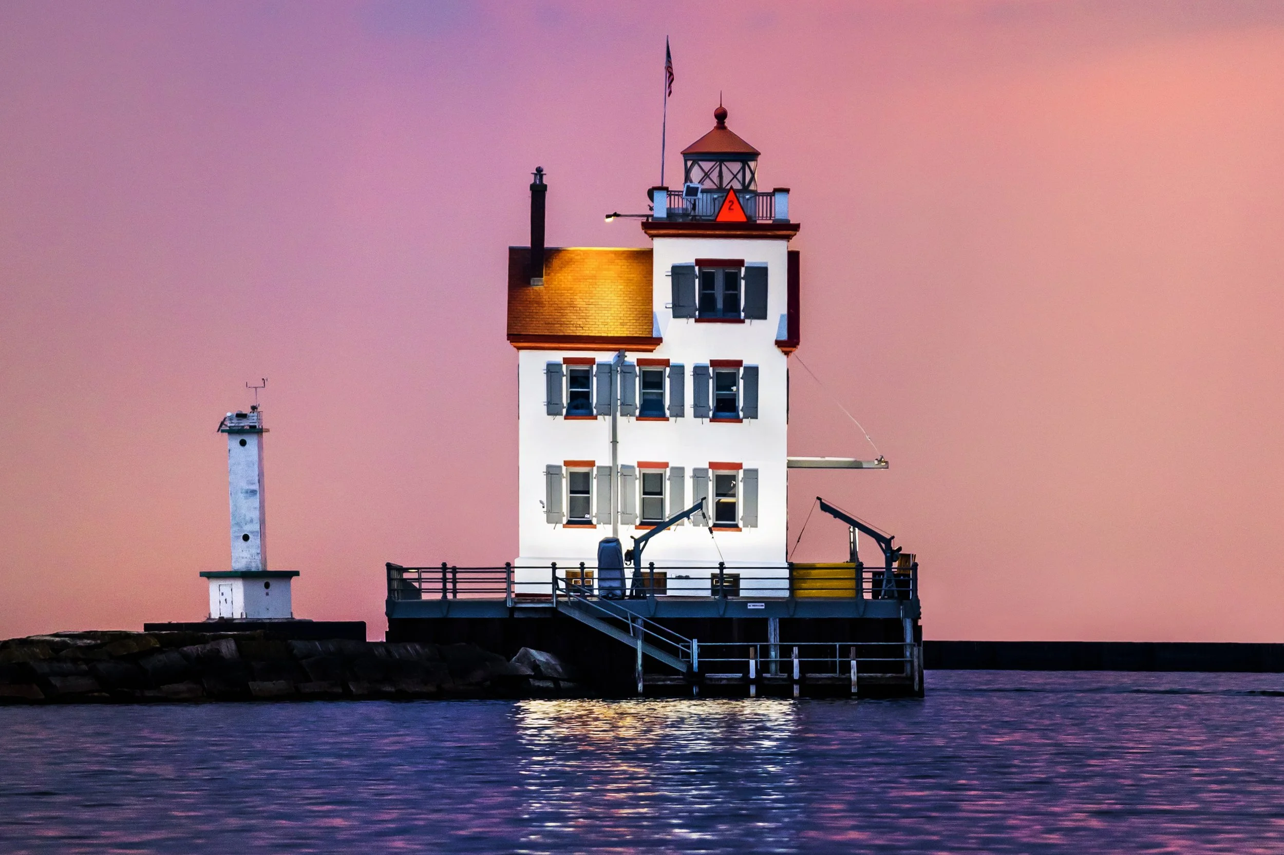 A lighthouse with a white structure and a smaller tower nearby, set against a pink and purple sunset sky, situated on a rocky pier extending into calm water.