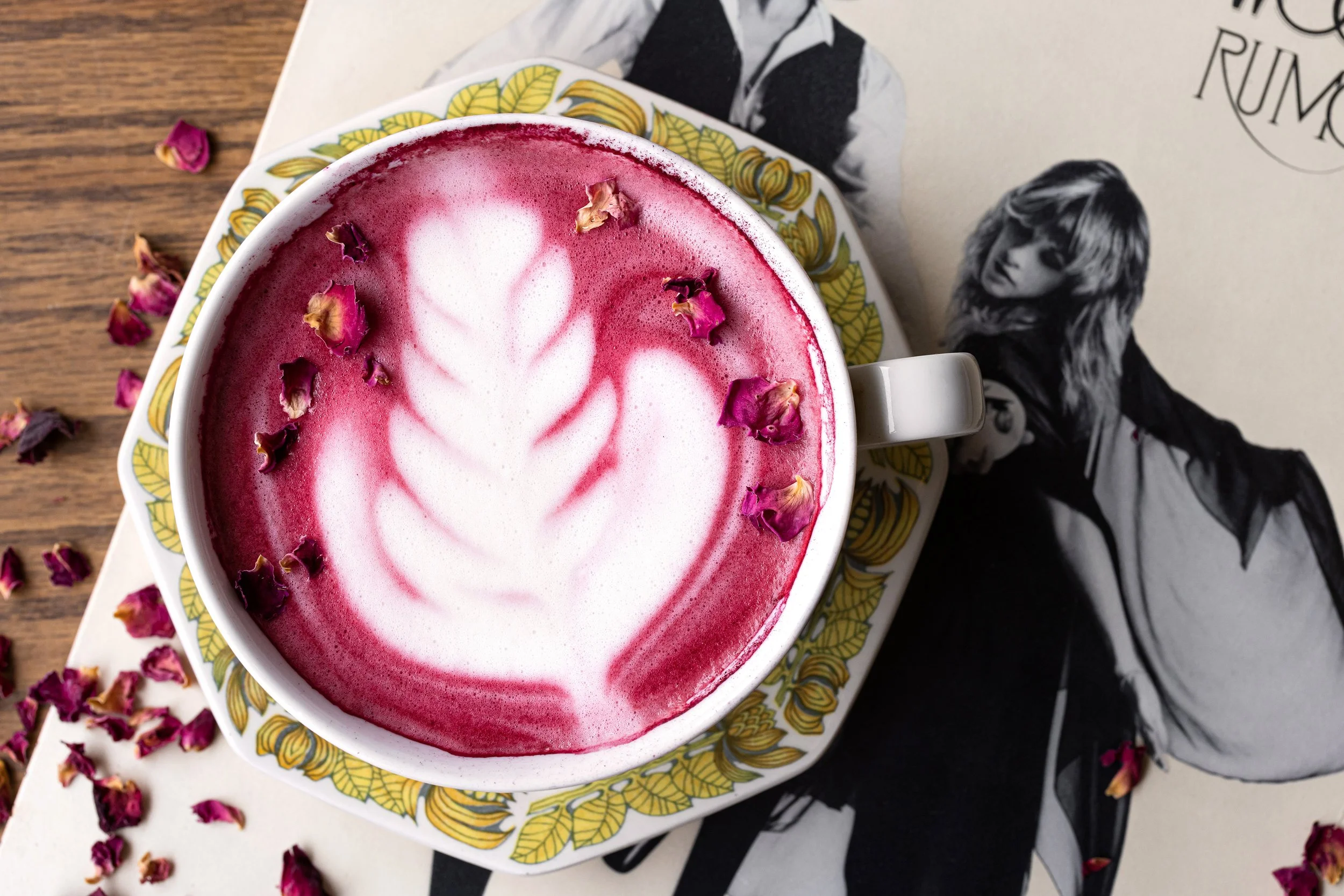 A cup of pink latte with foam art and dried rose petals on top, placed on a patterned saucer on a wooden table beside a black-and-white magazine featuring a woman with long hair.