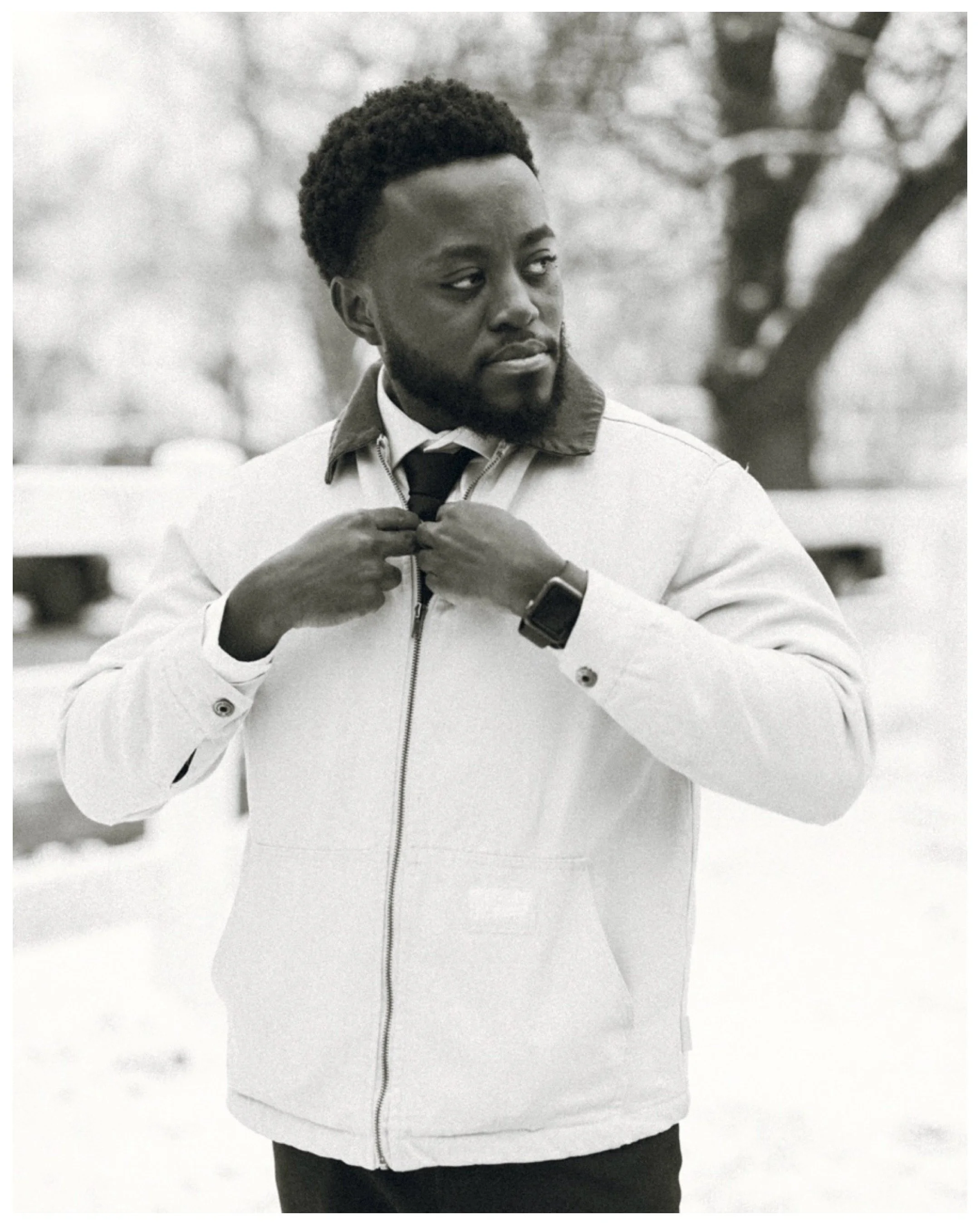 A man standing outdoors, adjusting a tie, wearing a light jacket, watch, and shirt with a collar, in a black and white photo.