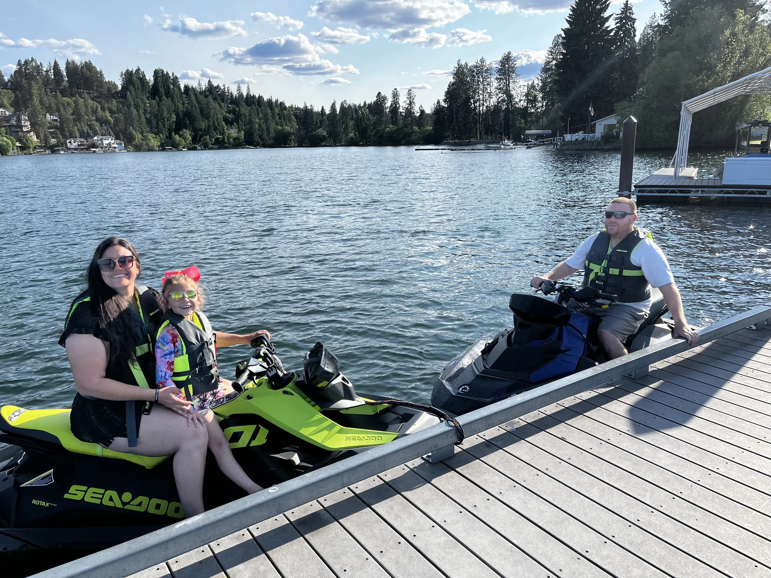Family sitting on jet skis at a lakeside dock with a boat and trees in the background on a sunny day.
