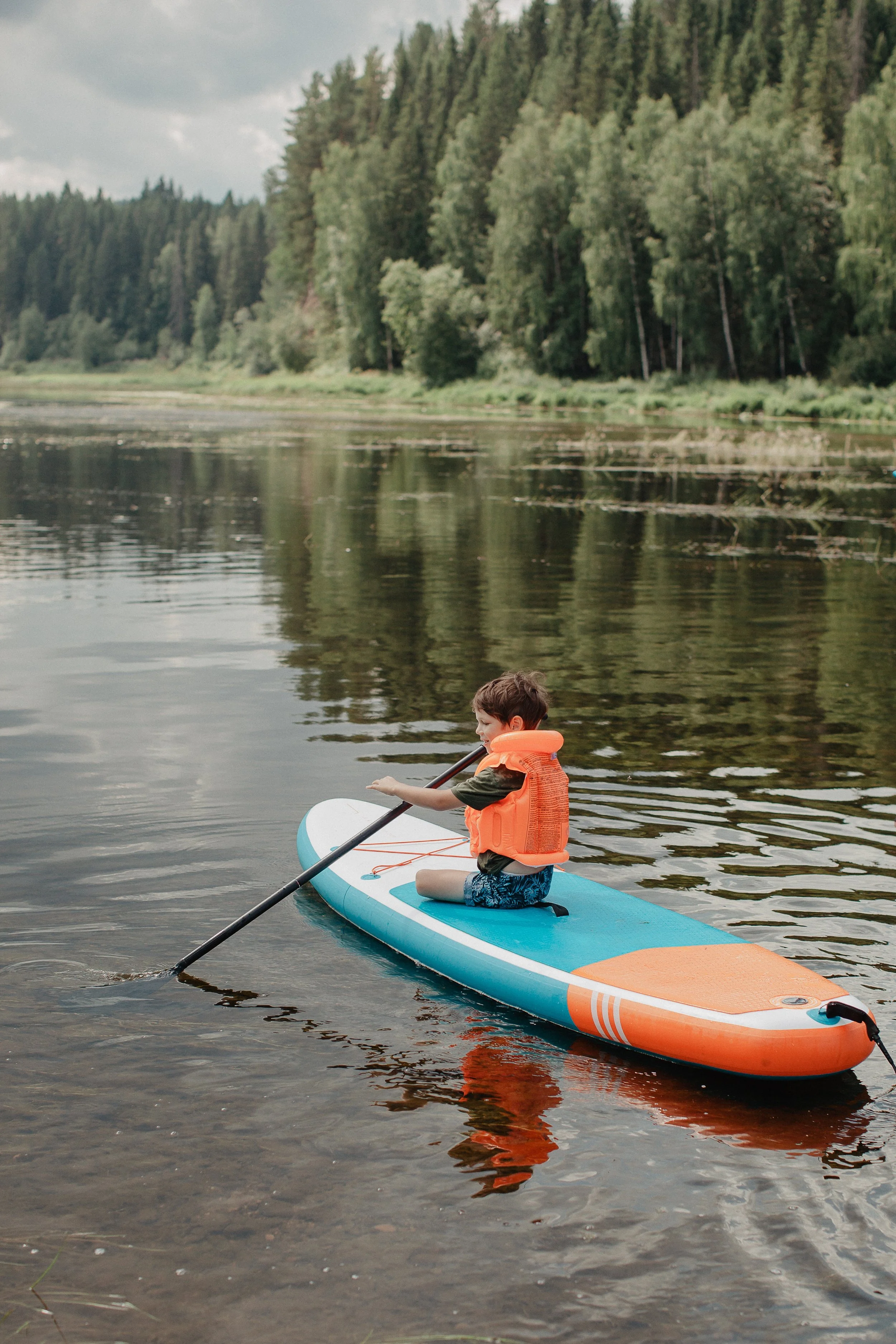 Child in an orange life vest kneeling on a blue paddleboard, paddling on a calm lake with trees and forest in the background.
