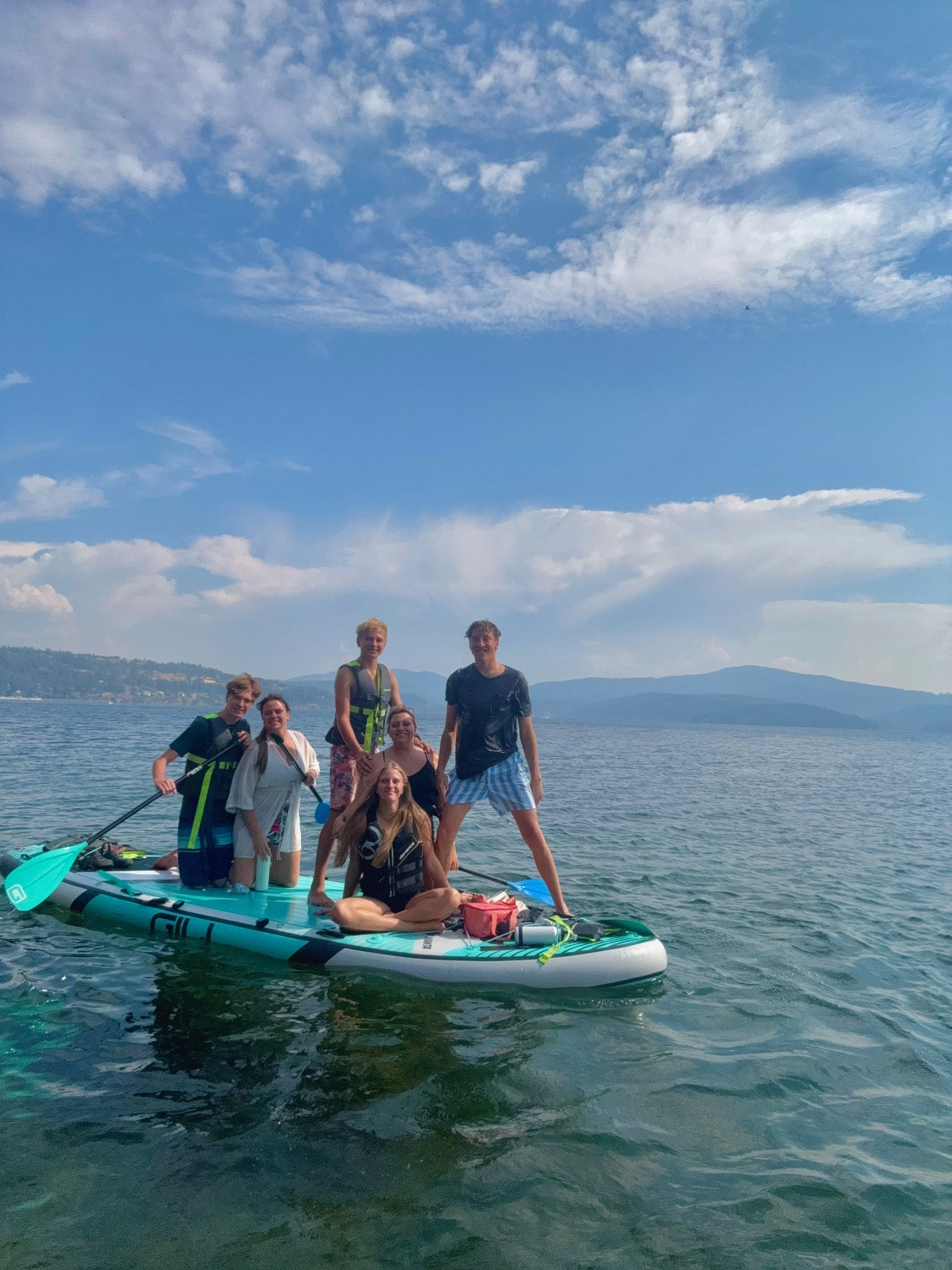 Group of seven young people standing and sitting on a paddleboard on a lake, with mountains and a blue sky with clouds in the background.