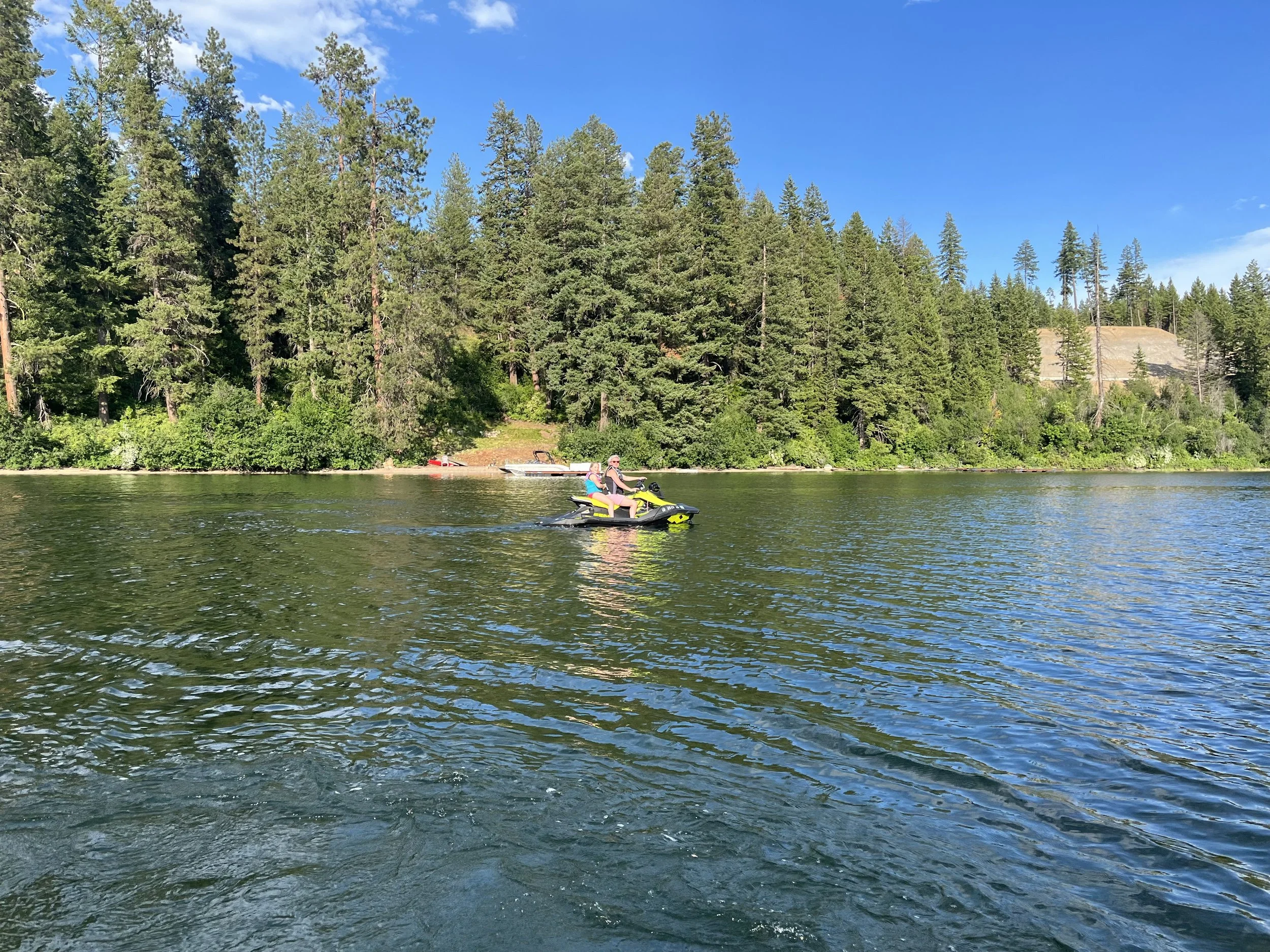 Two people riding a yellow jet ski on a lake, with a forested shoreline and a clear blue sky in the background.
