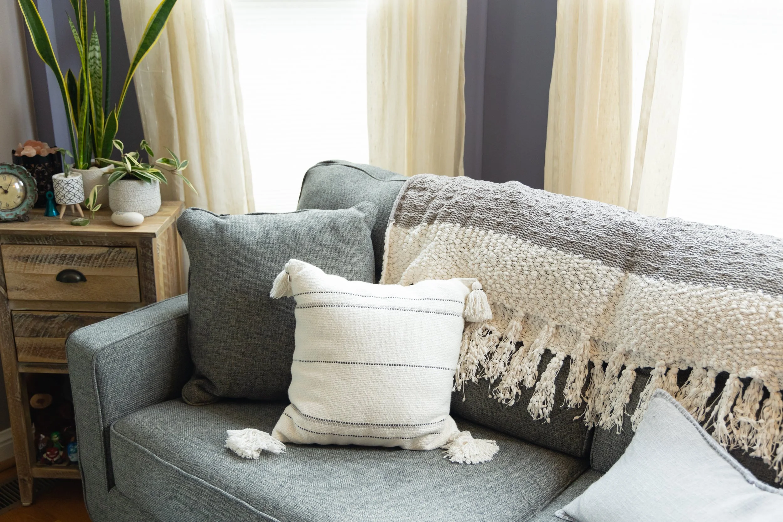 A view of a couch with throw pillows and a blanket, a wooden side table and curtains on the windows in an Empowered Spaces office.
