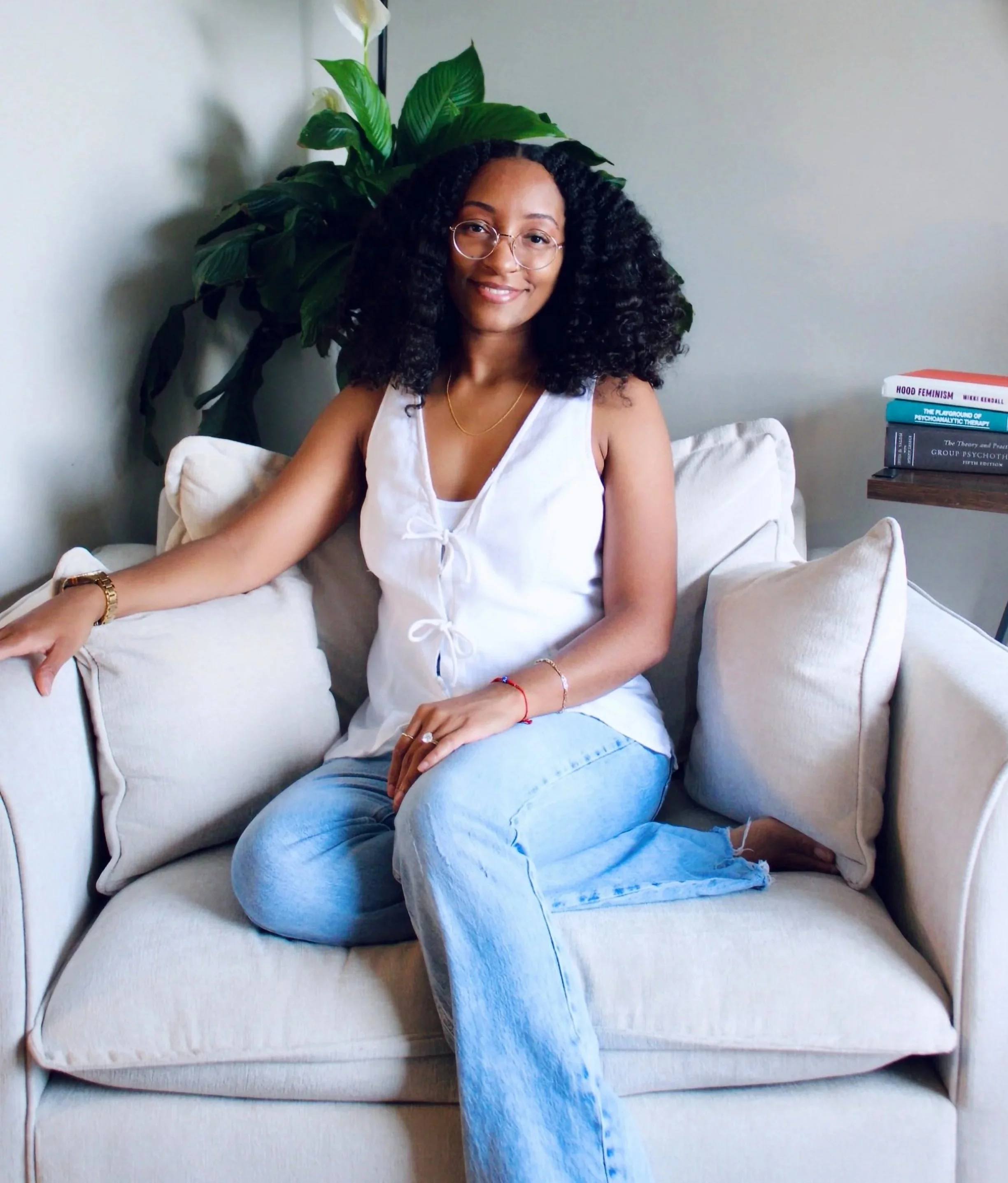 A woman smiling outdoors amid lush green foliage, wearing glasses, a black long-sleeve shirt, jeans, and a watch.