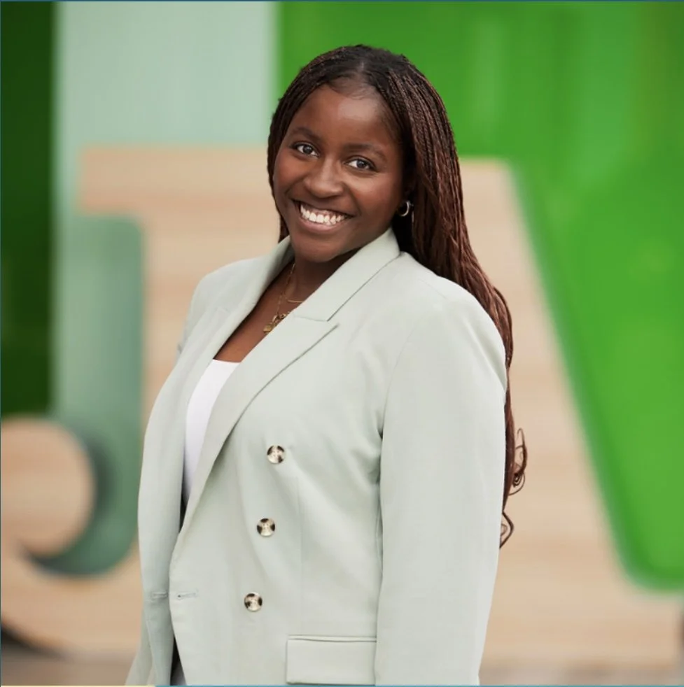 A smiling woman with long braided hair wearing a light-colored blazer and a white top, standing against a colorful background with green and beige tones.