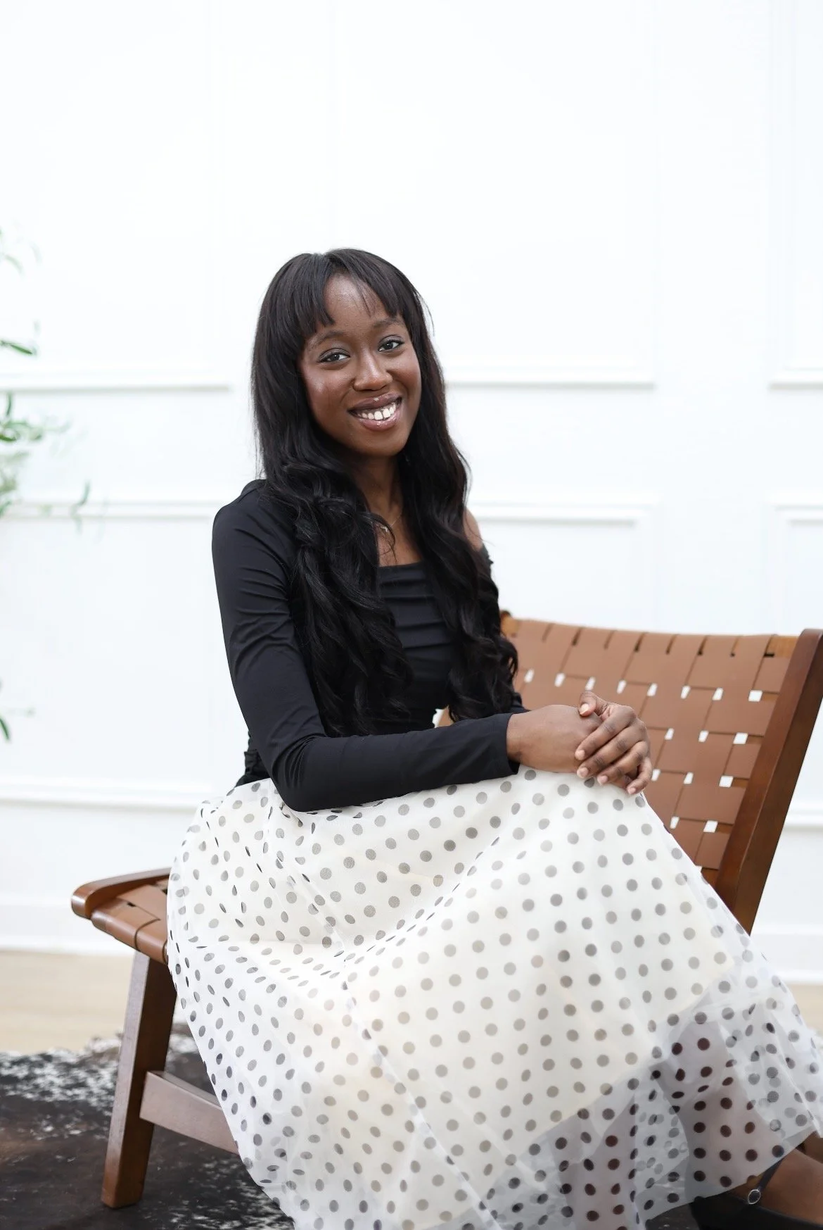 Lilianne Asokwah: A woman sitting on a wooden bench with a white wall background, wearing a black top and a white skirt with black polka dots, smiling at the camera.
