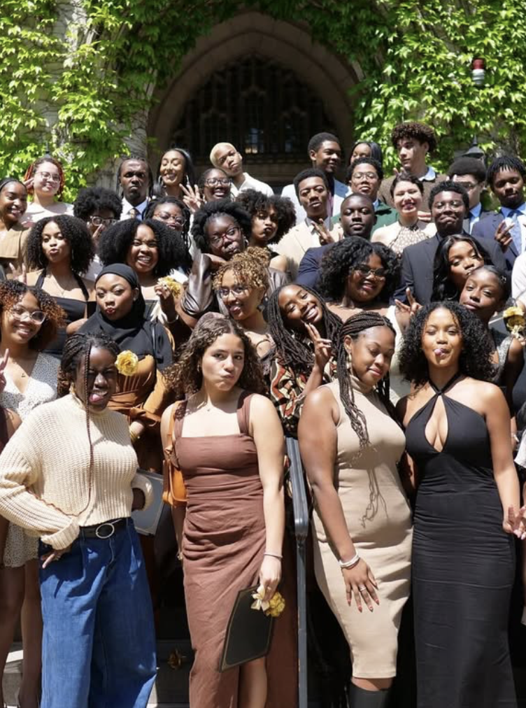 A large group of diverse young adults posing for a photo outside a building with greenery and an arched entrance, dressed in formal and semi-formal attire, smiling and making various hand gestures.