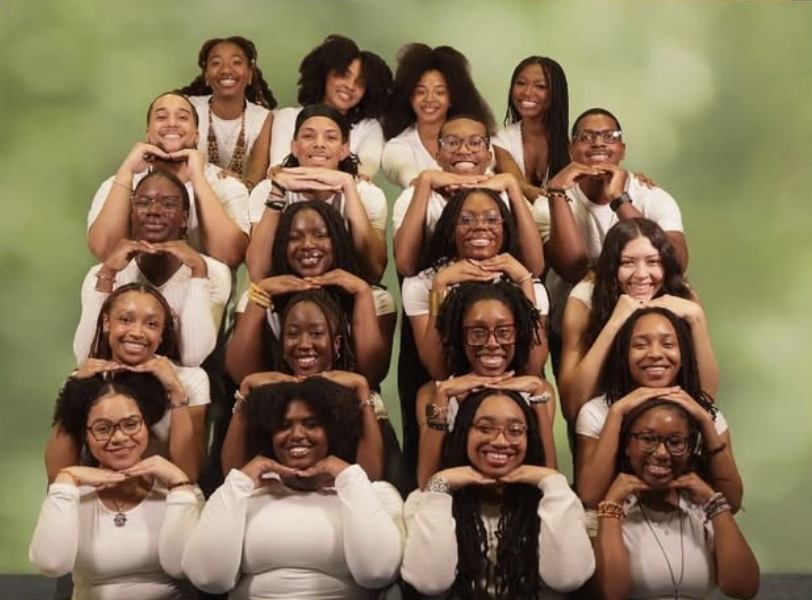 Group of young women and young men smiling and posing with their hands under their chins against a green background.