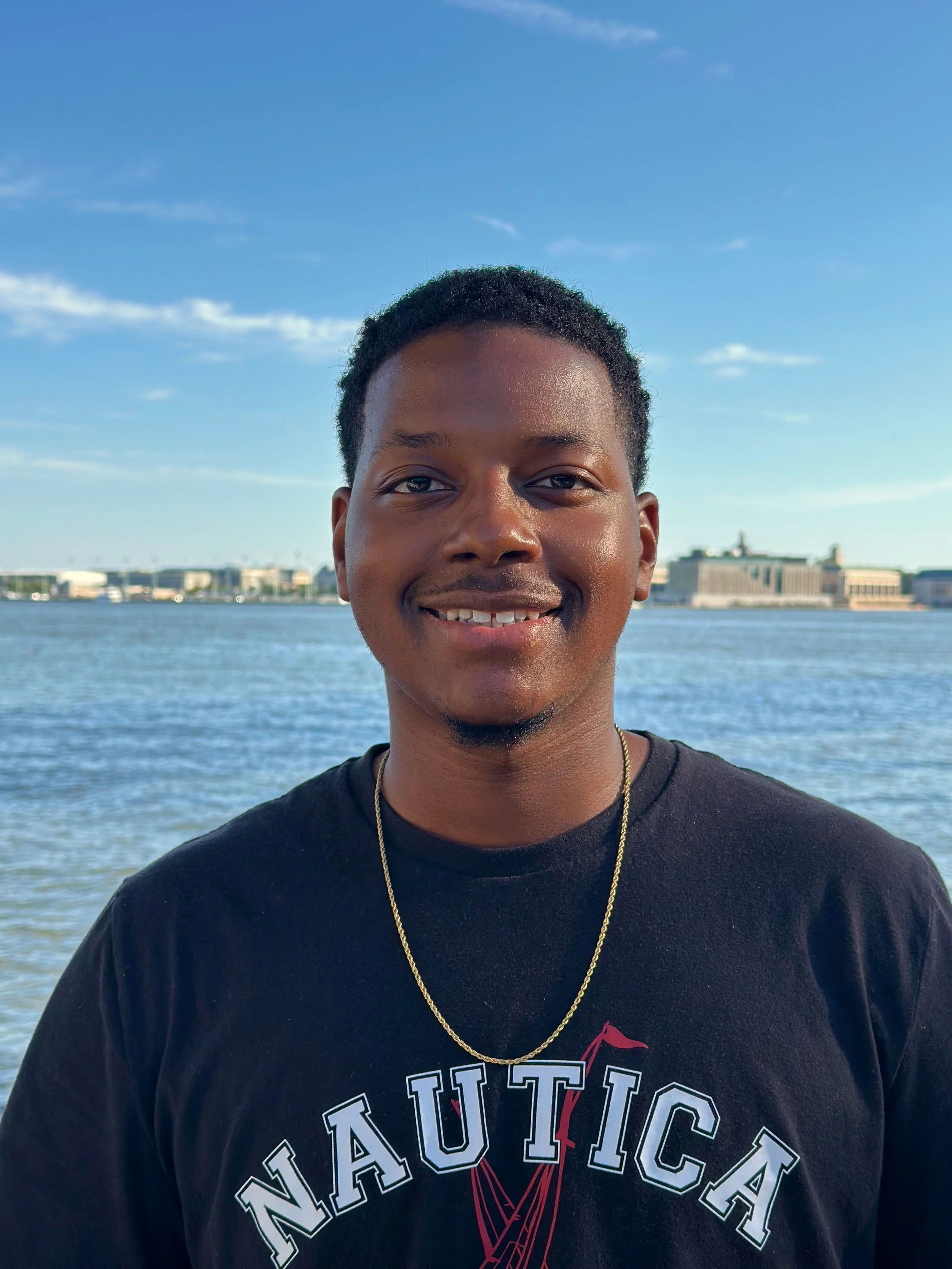 Akil Leggett, Jr: Smiling young man with short hair standing outdoors near water with buildings in the background during daytime.