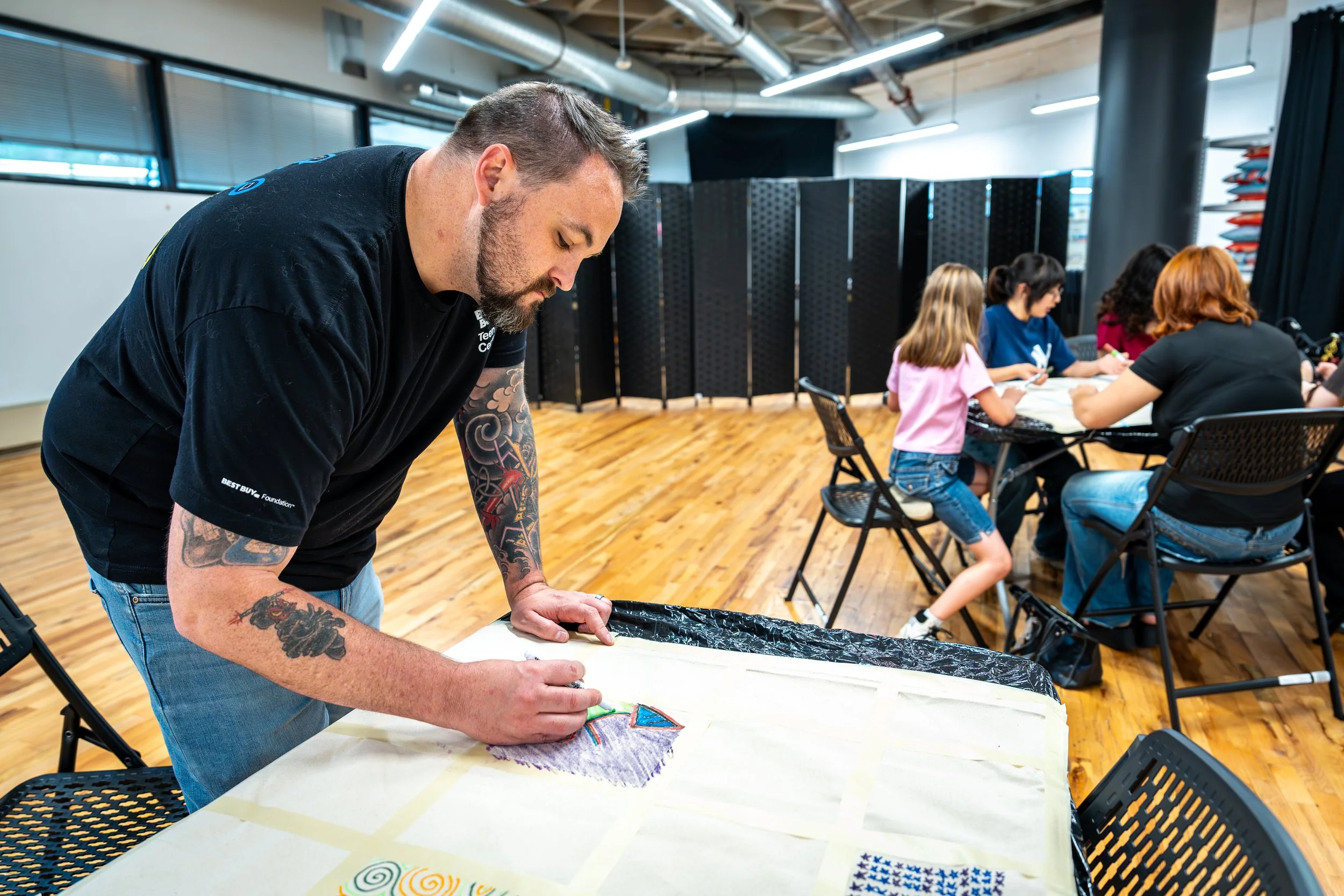 Man with tattoos drawing on fabric at a workshop, with children sitting at a table in the background.