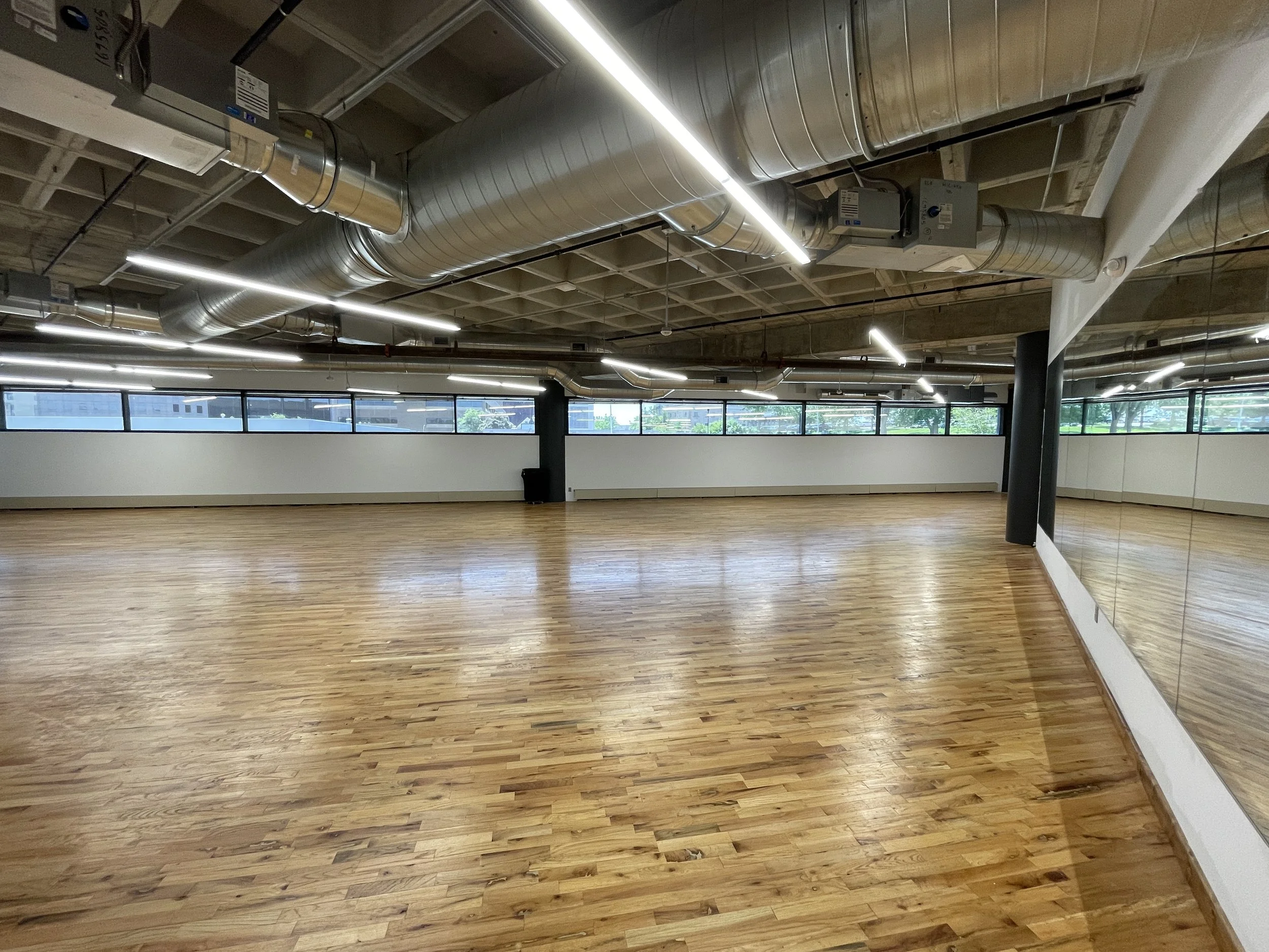 Empty dance or fitness studio with wooden floor, large wall mirror, and windows letting in natural light. Exposed ceiling with ducts and linear lighting.