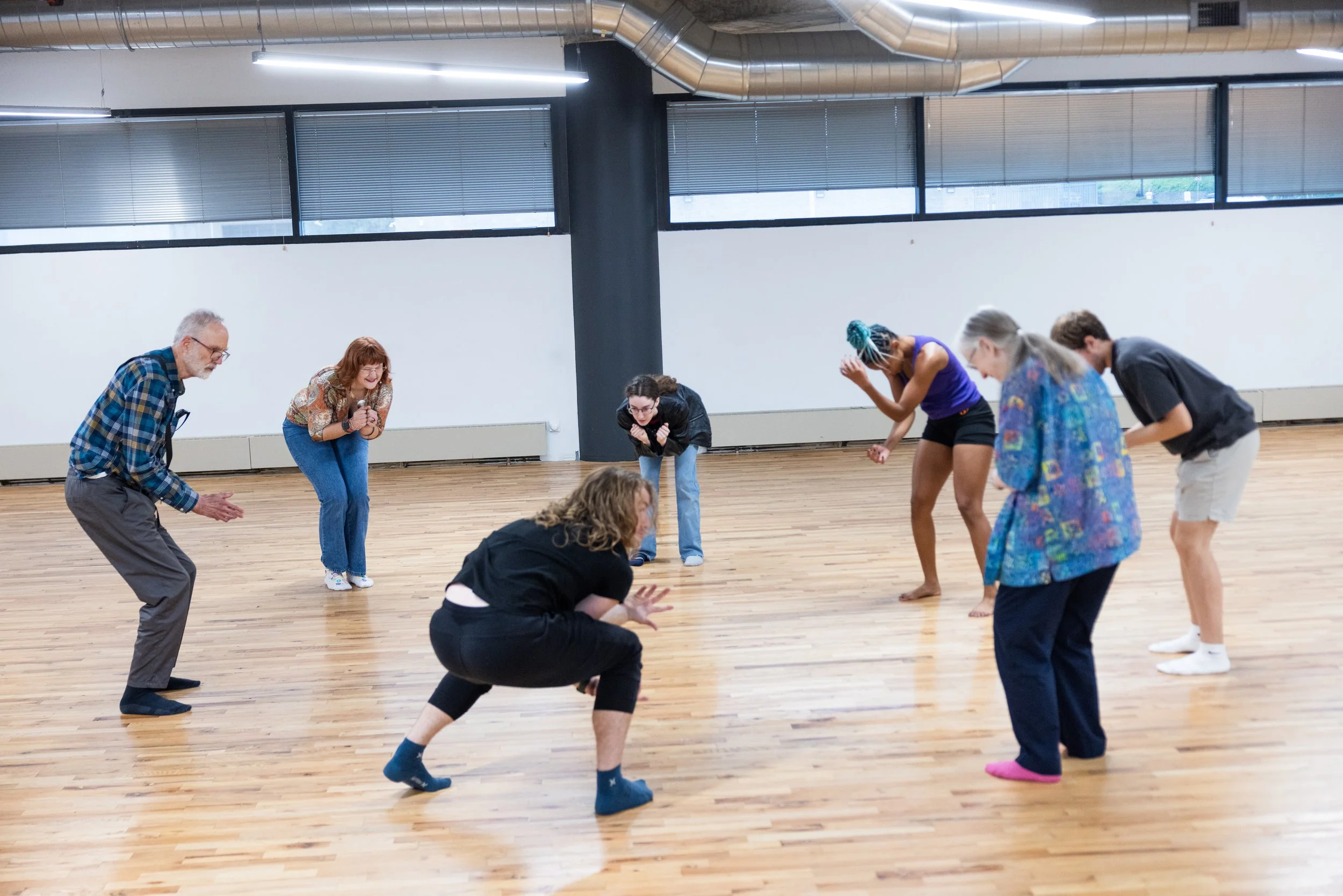 Group of eight people, including both men and women of diverse ethnicities, participating in a dance or movement class in a spacious studio with wooden floors, large windows, and industrial-style ceiling vents.