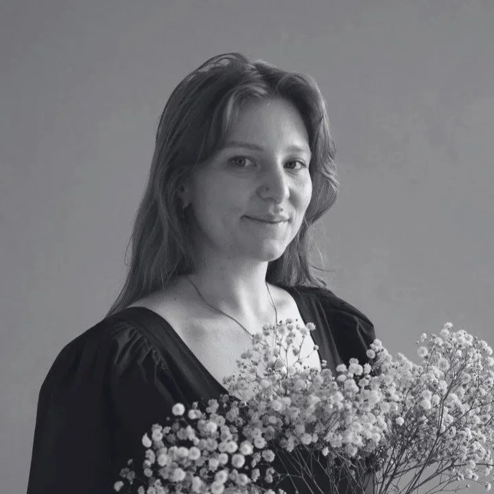 A woman with short hair and a septum piercing, smiling at the camera, wearing a sleeveless black top and a beaded necklace, in a room with posters, a lamp, and various signs on the wall.