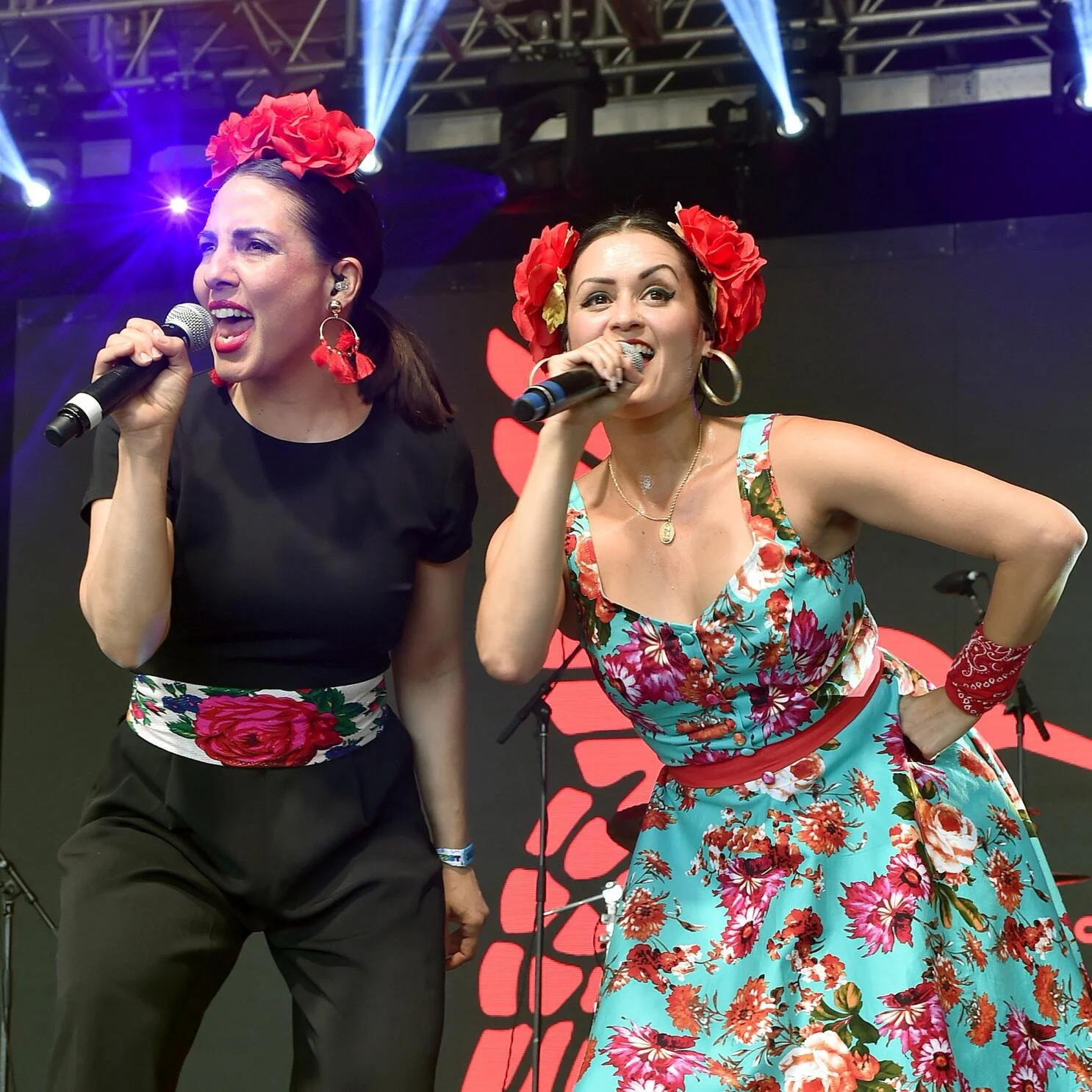 Two women singing into microphones on stage. The woman on the left wears a black outfit with floral accents, while the woman on the right wears a blue floral dress. Both have red flowers in their hair.