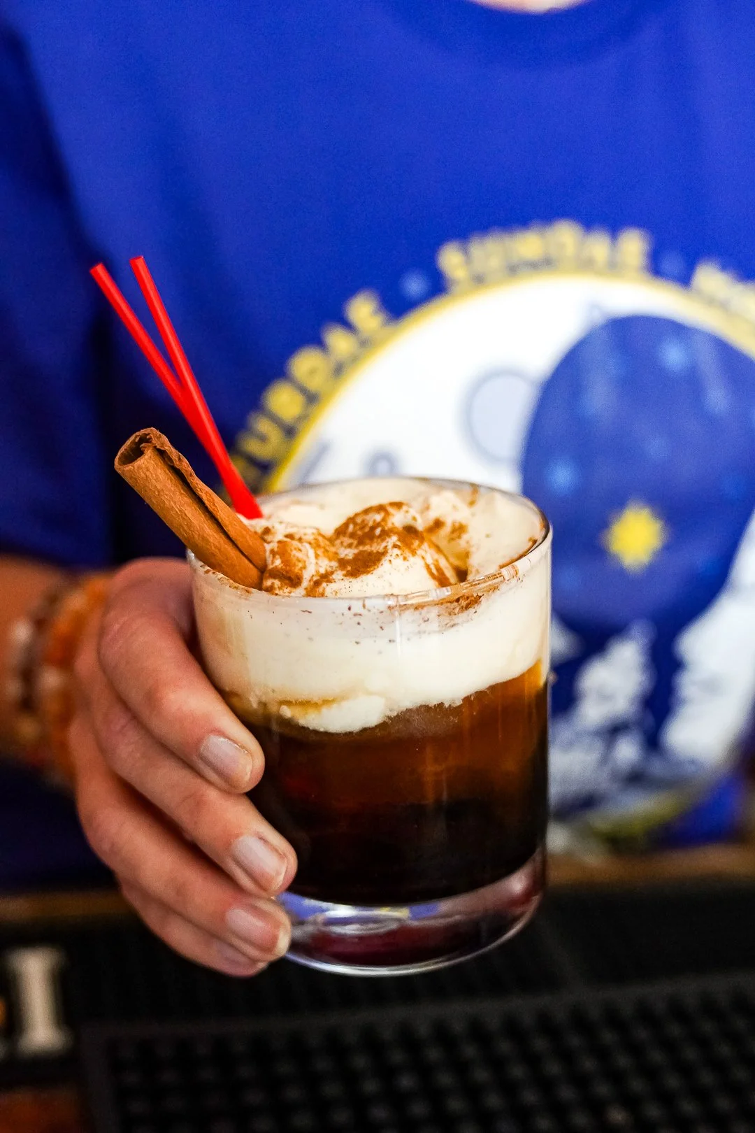 A hand holding a glass of dark iced cocktail topped with whipped cream, dusted with cinnamon, and garnished with a cinnamon stick. Two red drink stirrers are placed in the glass.