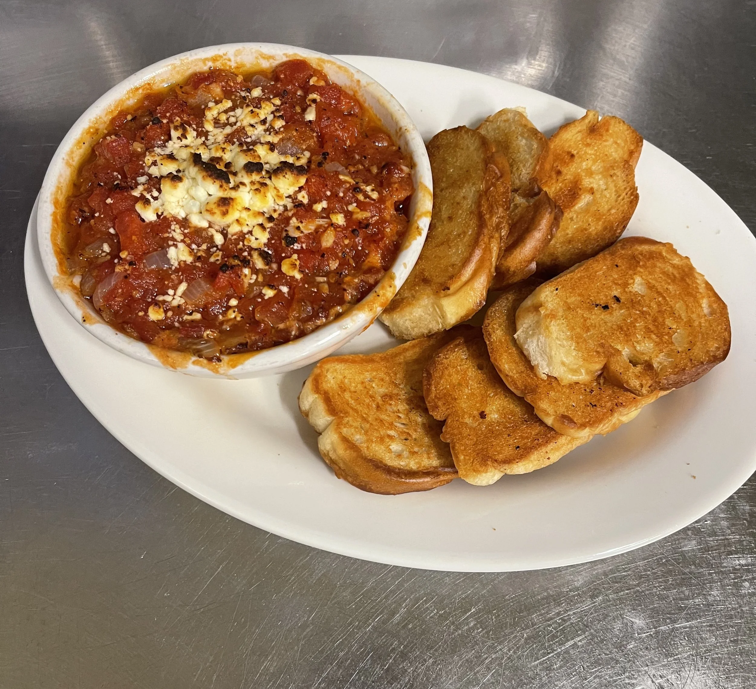  A white plate holds a bowl of red dip, likely salsa or bruschetta, topped with cheese and herbs. The dip is accompanied by toasted bread slices on the right side of the plate, set against a stainless steel background.  Meta AI Response: A white plat
