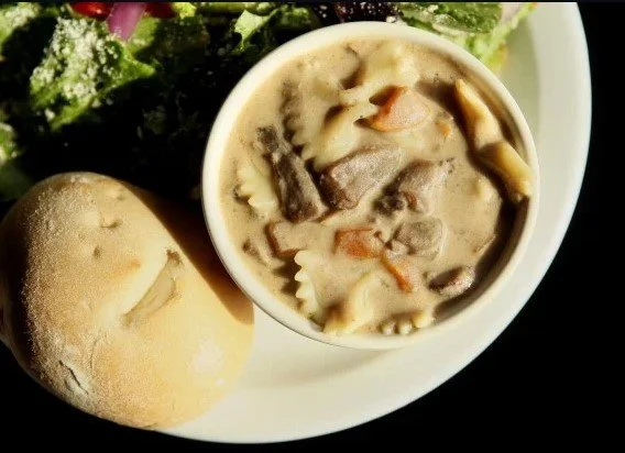 A bowl of beef stroganoff soup sits on plate next to a roll and some salad.
