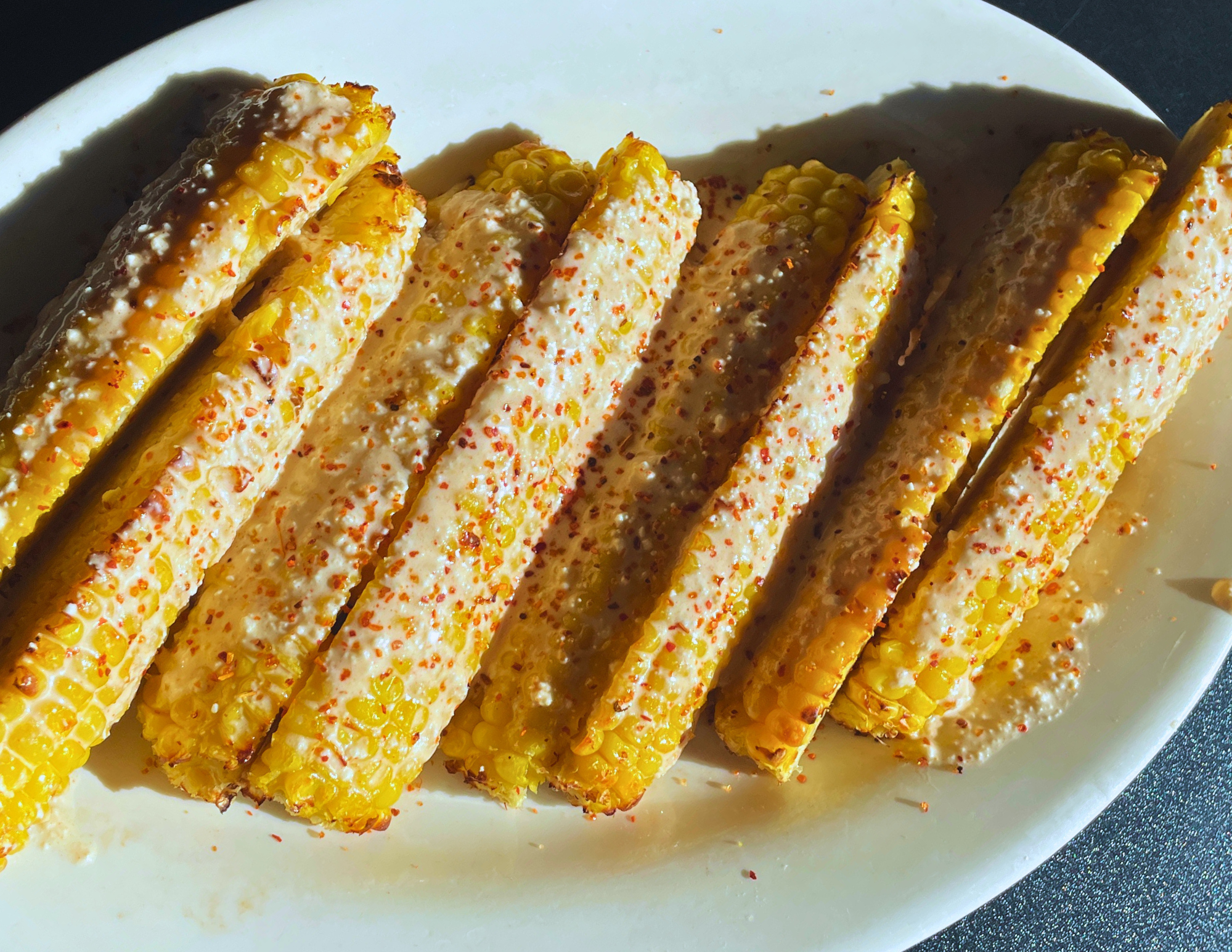 Six grilled corn ribs on a white plate, coated with a creamy white sauce, sprinkled with red chili powder and grated cheese, served as Mexican-style elote ribs.